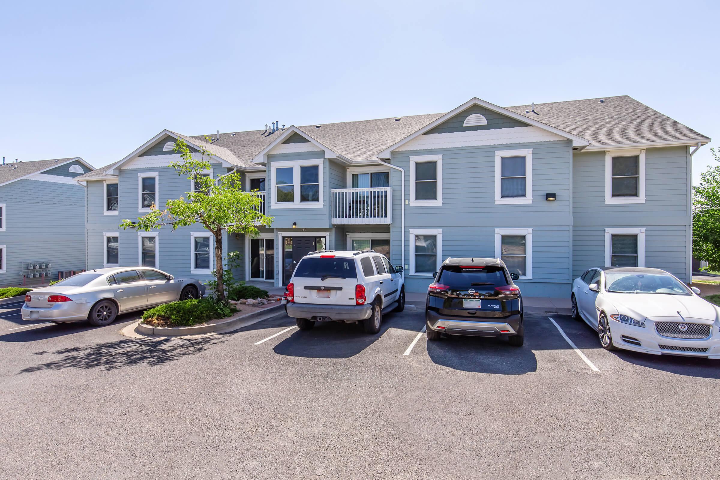 A two-story light blue apartment building with several parked cars in front. The building features multiple windows, a balcony on the second floor, and a small tree in the landscaping. The scene is set on a clear, sunny day.