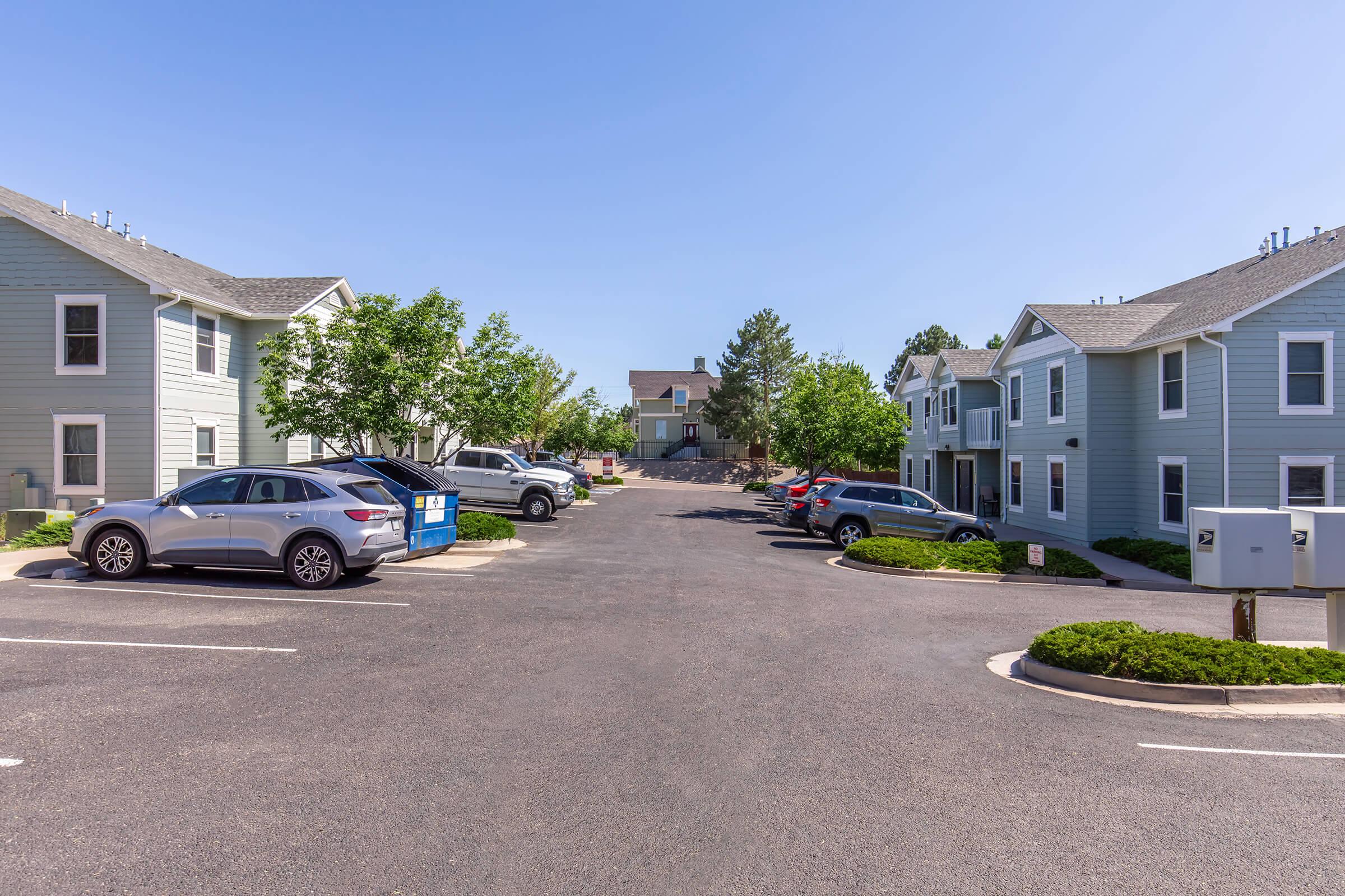 A view of a residential parking lot with several parked cars, including a gray SUV and a blue dumpster, surrounded by light blue apartment buildings. The scene is under clear blue skies, with green trees lining the sides of the lot, creating a peaceful, suburban atmosphere.