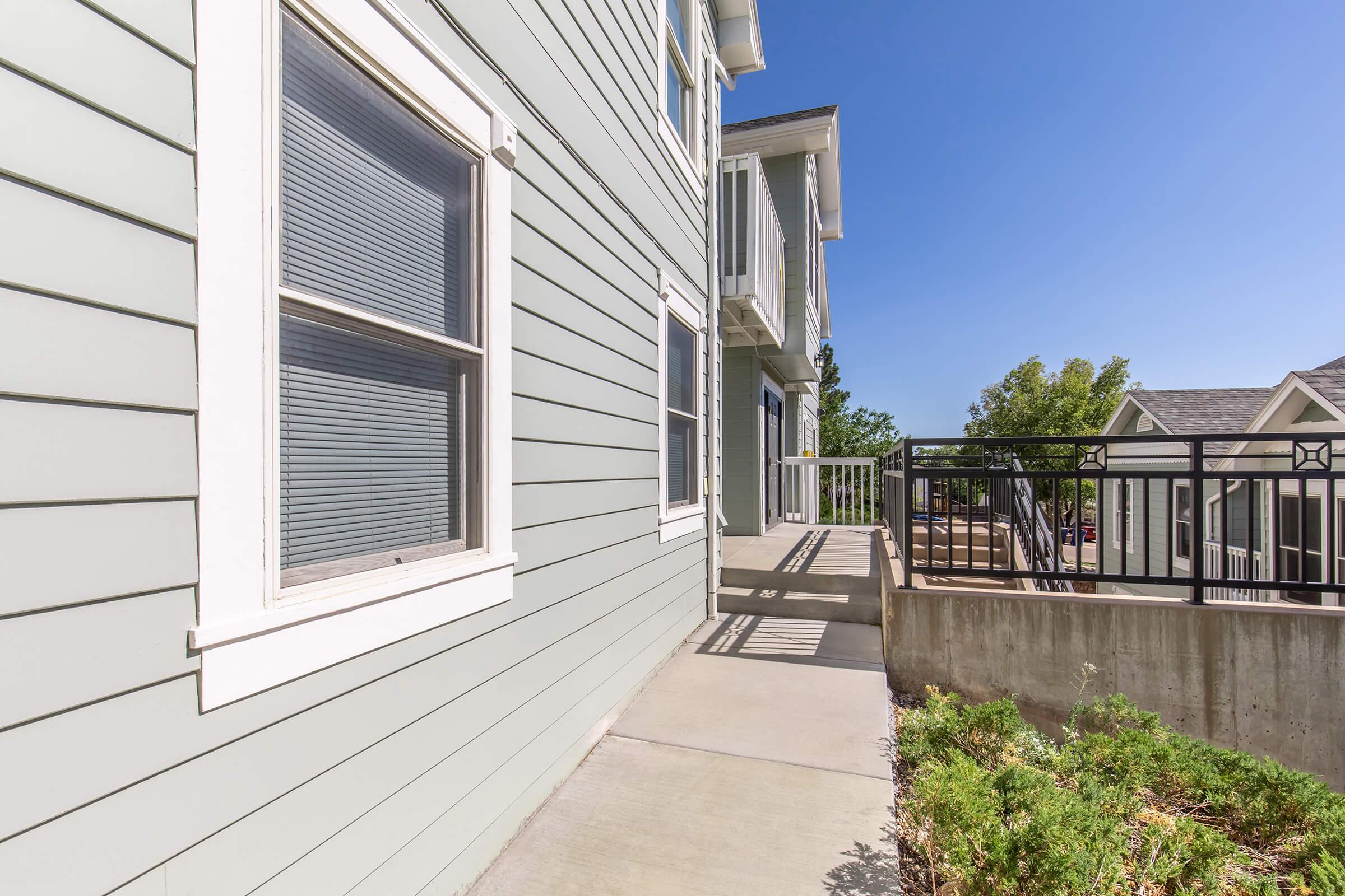 A pathway leading alongside a light blue house, with large windows and white trim. The surroundings feature a concrete area with greenery, and clear blue skies above. The image captures the modern design of the building and the outdoor space, emphasizing a well-maintained environment.