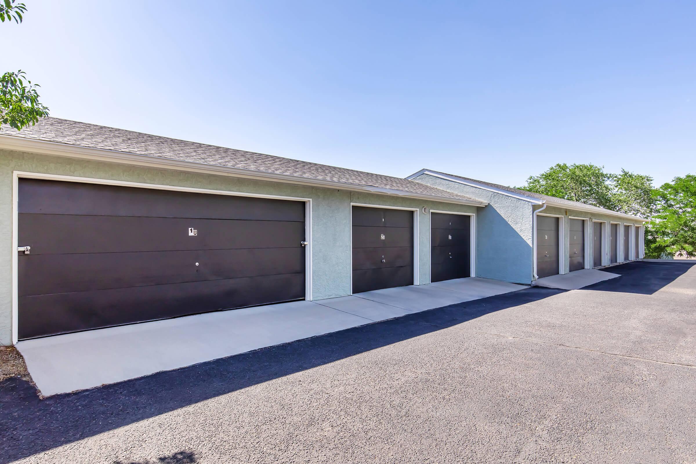 Row of garage doors with a smooth concrete driveway, set against a clear blue sky. The garages feature black doors, some with numbers, and are part of a light blue building. Green trees are visible in the background, adding a touch of nature to the scene.