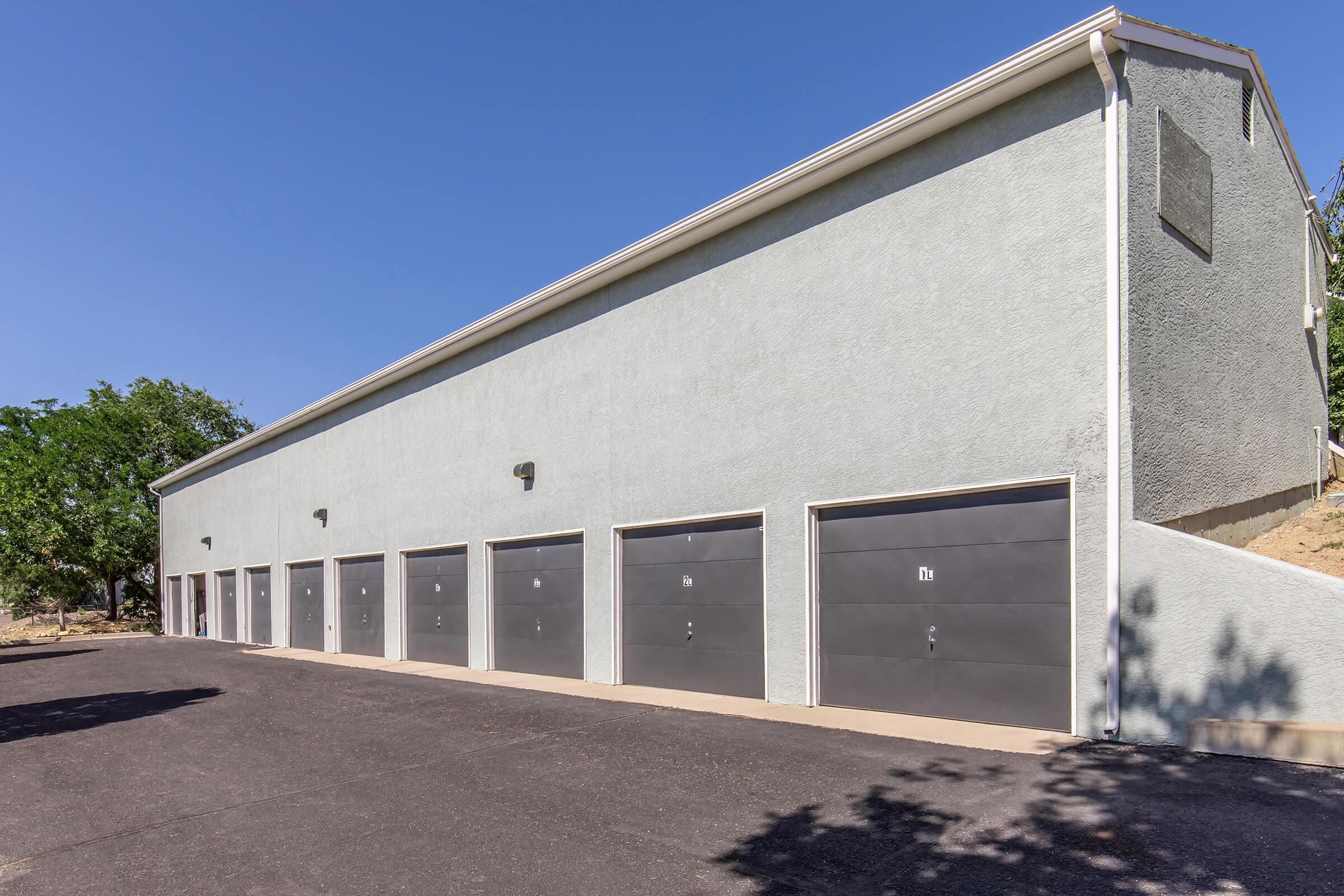 A row of gray garage doors set in a light-colored building, situated on a paved driveway. The structure features a simple design, with large windows and exterior lighting. Green trees are visible in the background under a clear blue sky. Shadow patterns are cast on the ground.