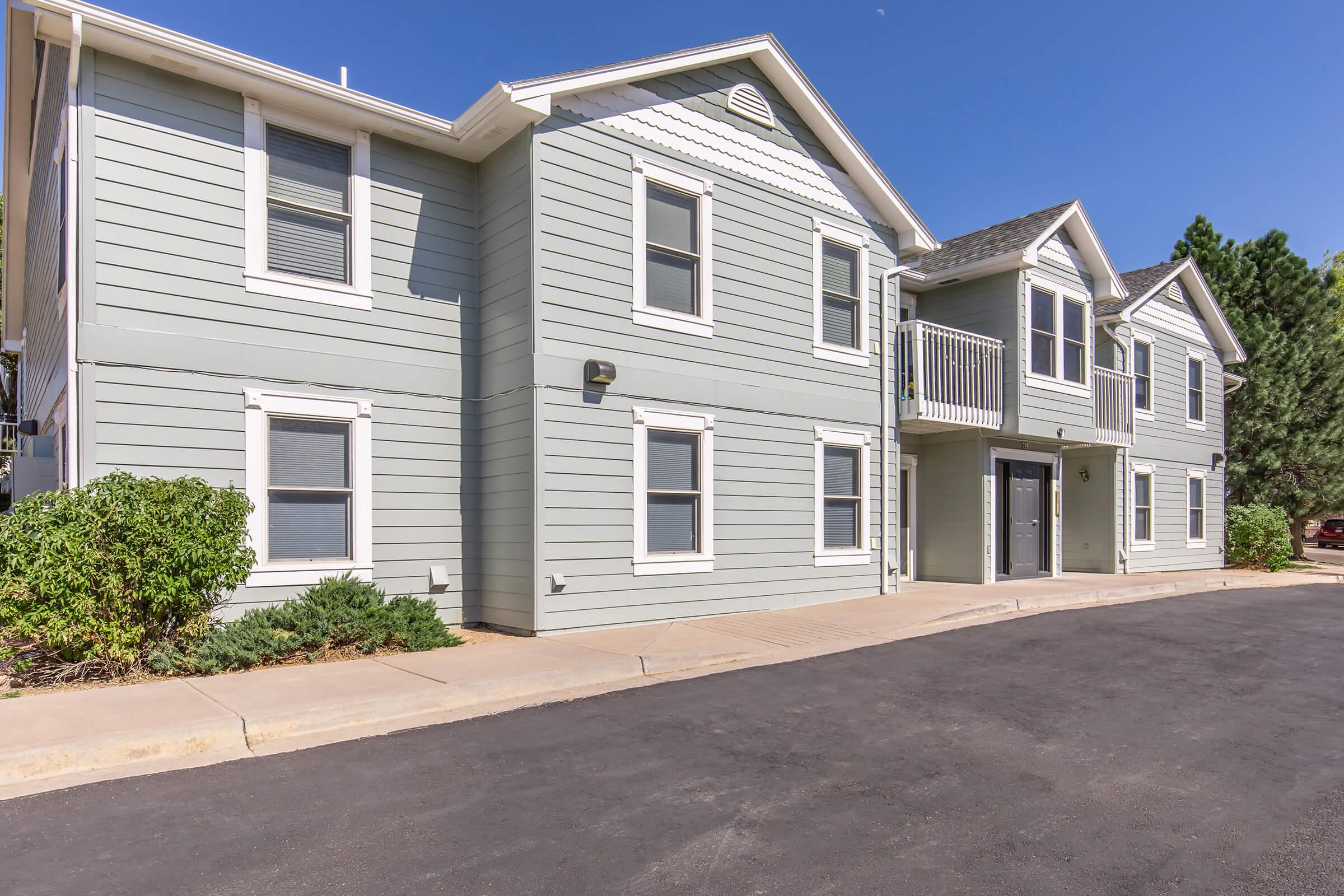 Exterior view of a light green multi-family building with a gray roof. The structure features multiple windows, a balcony, and a front walkway. A neatly maintained landscape with shrubs is visible along the perimeter. The scene is set on a clear blue sky day, highlighting the building's modern design.