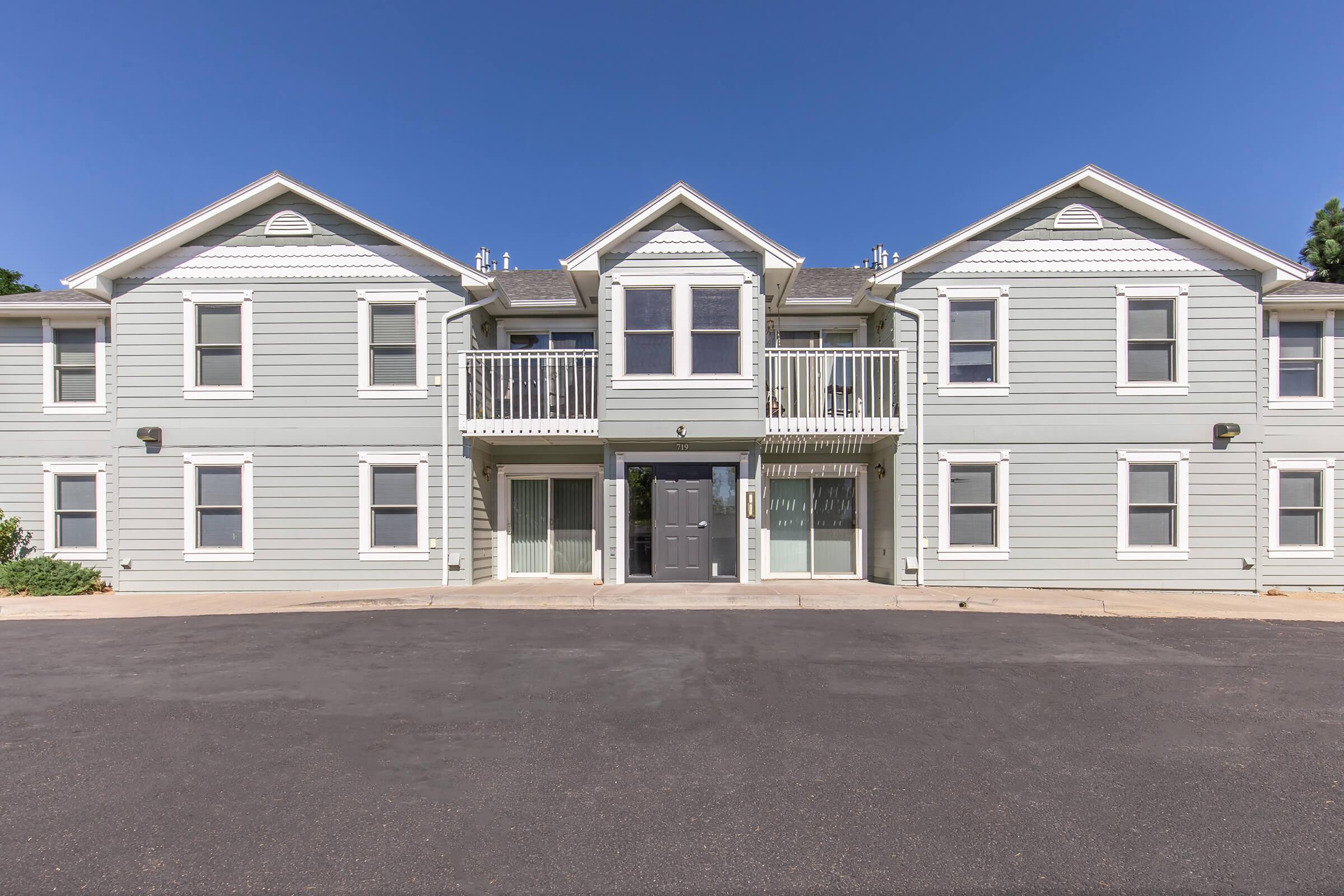 A two-story residential building with light gray siding, multiple balconies, and large windows. The entrance features a double door, and the surrounding area has a paved driveway. The sky is clear and blue, enhancing the modern appearance of the building.