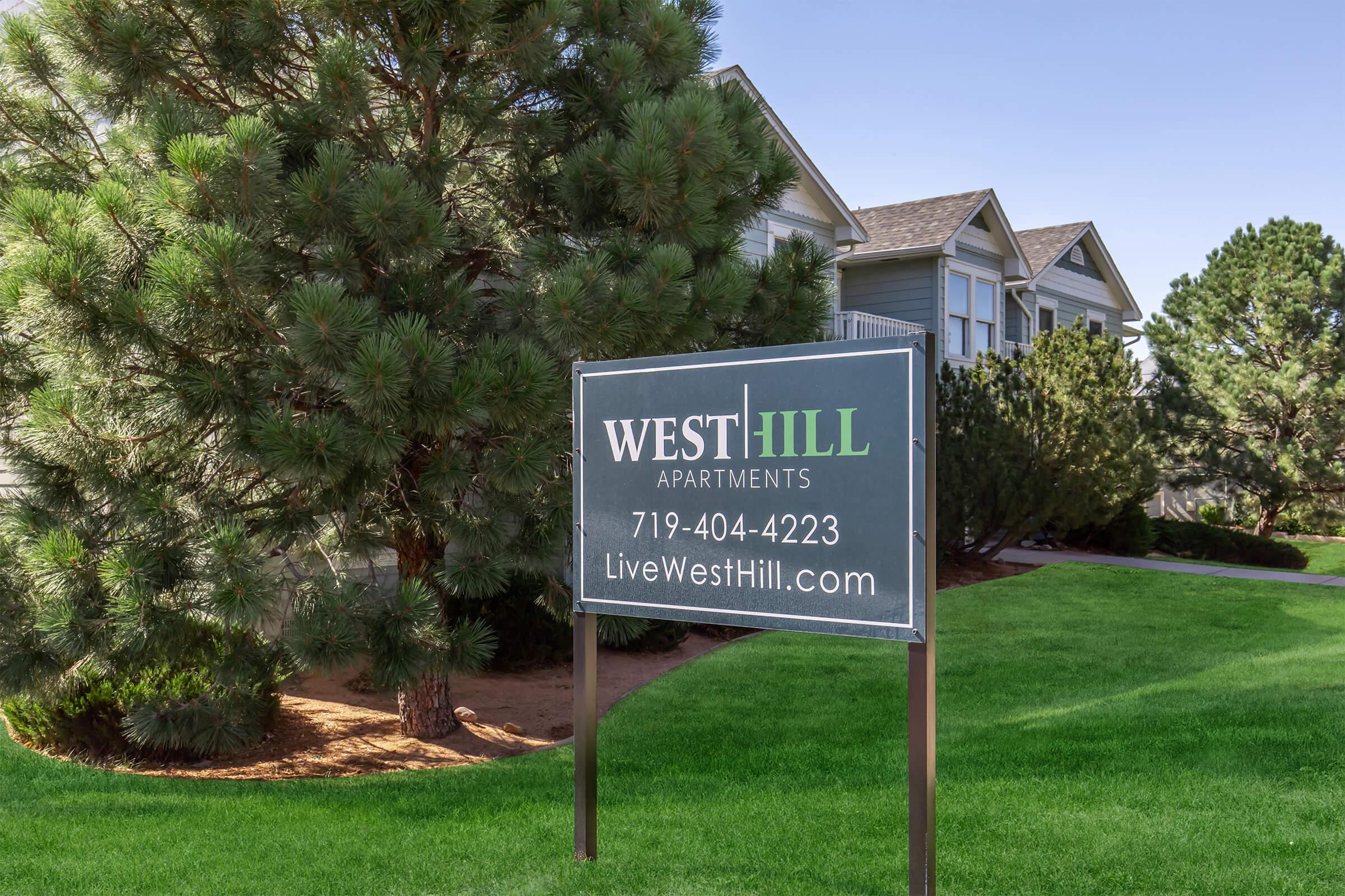 Sign for West Hill Apartments, featuring the contact number 719-404-4223 and the website LiveWestHill.com. The sign is situated in a landscaped area with greenery, offering a view of nearby residential buildings.