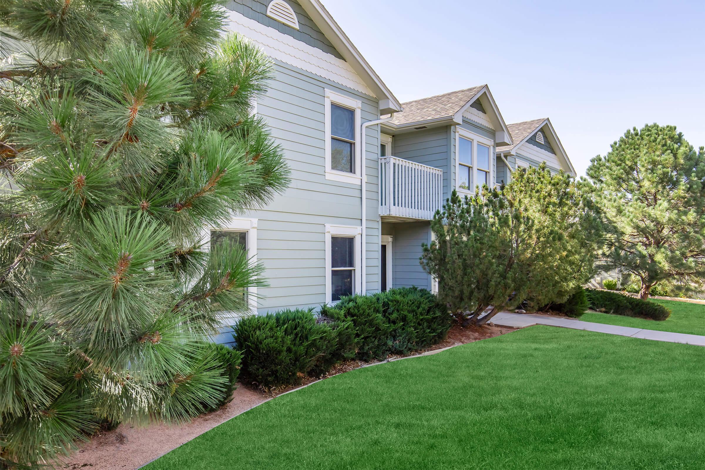 A well-maintained two-story residential building with light blue siding. The foreground features lush green grass and landscaped shrubs, while tall pine trees provide a natural backdrop. A small balcony is visible on the upper level, enhancing the building's charm. The clear sky suggests a sunny day.