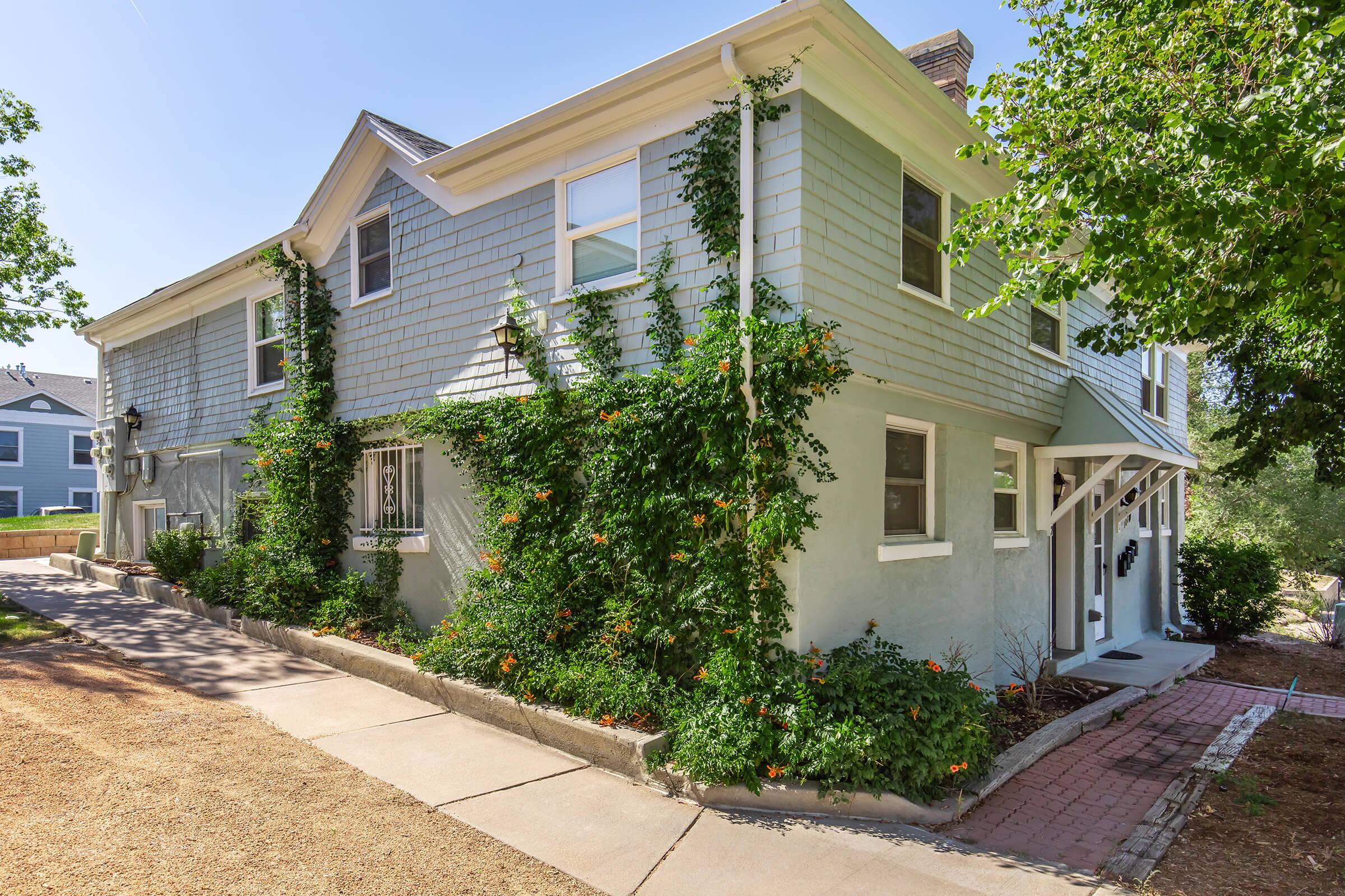 A two-story house featuring light blue siding, large windows, and covered entryways. Ivy and orange flowers climb the walls, and a pathway leads to the entrance. The surrounding area is landscaped with gravel and greenery, giving the property a welcoming and well-maintained appearance.