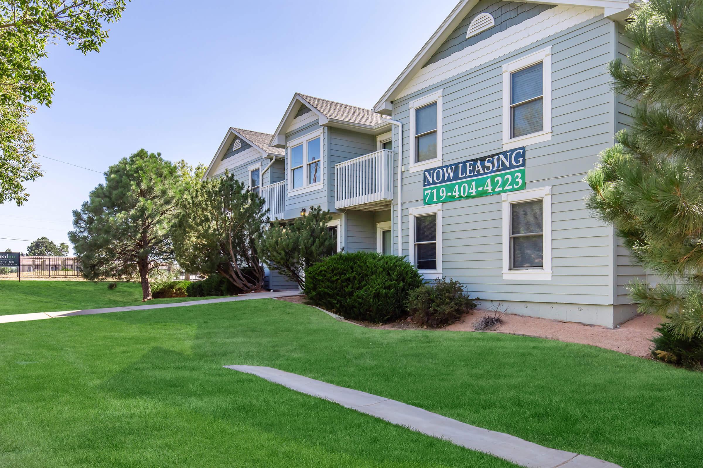 A multi-unit residential building with a light blue exterior and white trim. The building features several windows and a balcony. In front, a green lawn is visible, along with landscaped shrubs and trees. A large sign stating "NOW LEASING" with a phone number is prominently displayed on the building.