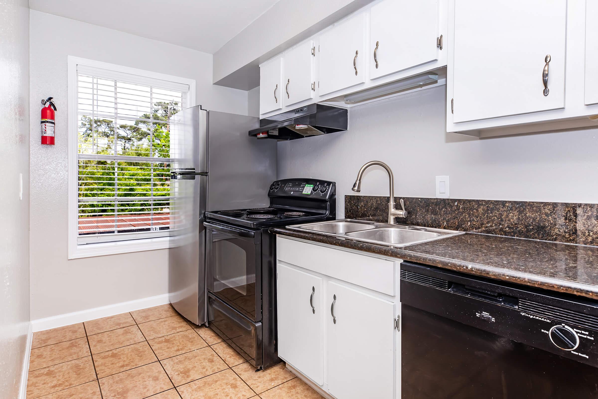 A modern kitchen featuring white cabinets, a stainless steel refrigerator, an electric range, and a double sink. The granite countertop contrasts with the light cabinetry. Natural light streams in from a window with blinds, and there is a fire extinguisher mounted on the wall. The floor is tiled with beige tiles.