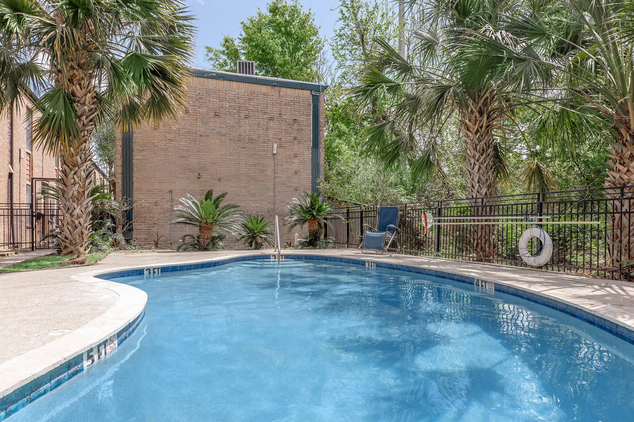 A serene swimming pool surrounded by lush greenery and palm trees. The pool is inviting, with clear blue water reflecting the sky. A lounge chair is placed nearby, and a life buoy hangs on the poolside. The scene is set against a brick building, enhancing the tranquil atmosphere.