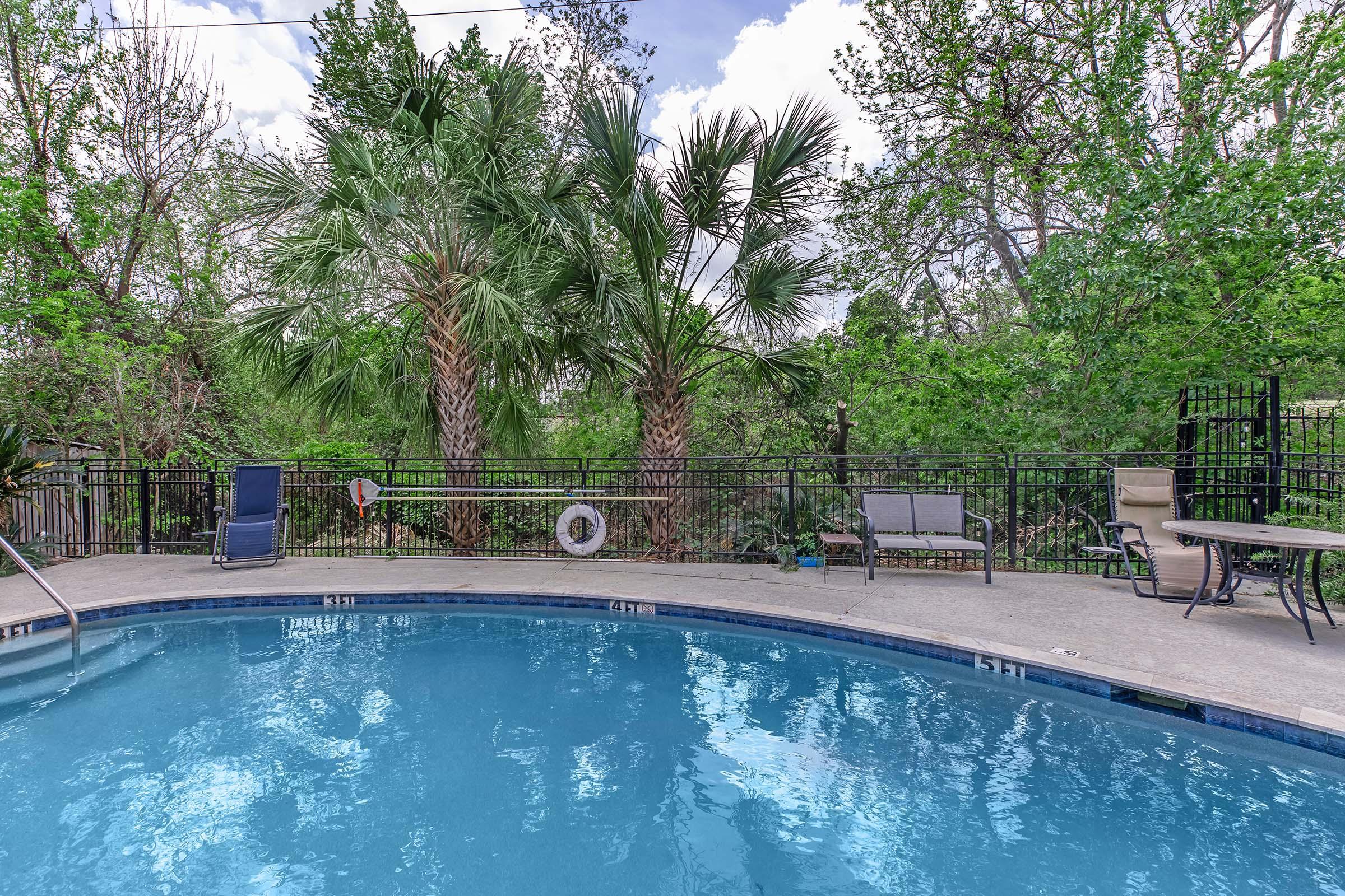 A tranquil outdoor pool area surrounded by lush greenery, featuring tall palm trees and a black metal fence. Several lounge chairs are placed by the poolside, with a table nearby. The clear blue water reflects the sky, creating a serene and inviting atmosphere for relaxation.