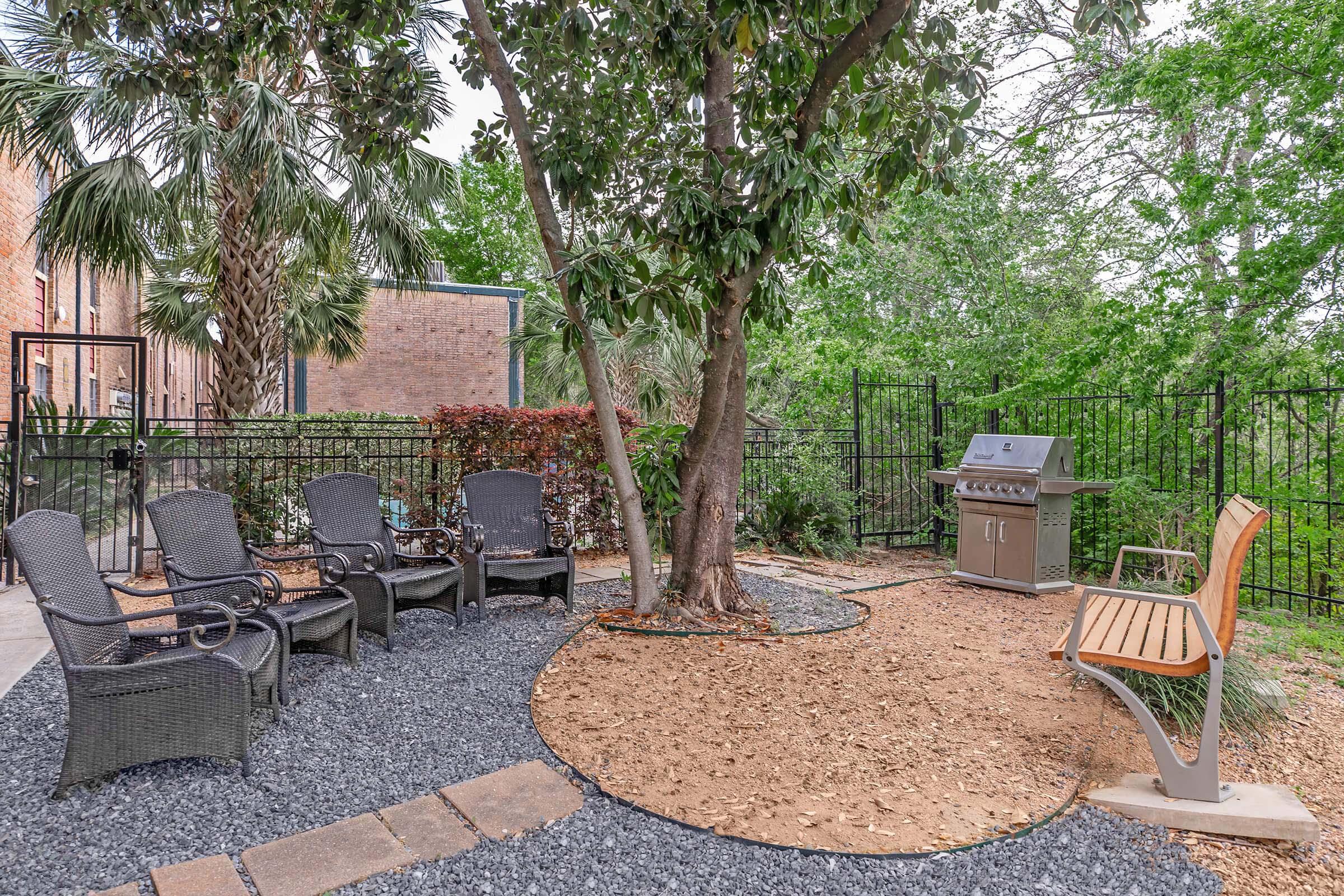 A cozy outdoor area featuring a set of four black wicker chairs arranged around a stone path, next to a bench. A grill stands nearby, surrounded by gravel and mulch. Lush green trees provide a natural backdrop, and a black fence encloses the space, creating a welcoming setting for relaxation or gatherings.