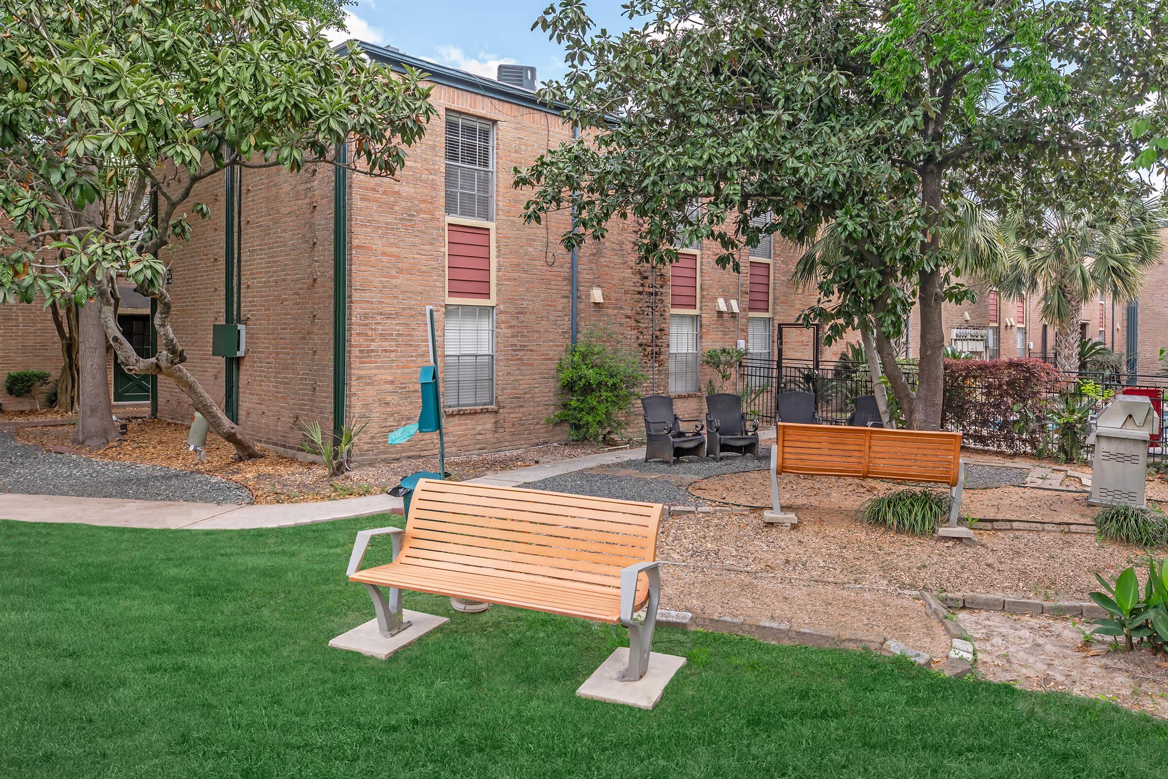 A peaceful outdoor area featuring a wooden bench on grassy ground, surrounded by trees and shrubs. In the background, there are brick buildings with windows, and a small seating area with chairs. The scene is well-maintained, offering a tranquil space for relaxation.