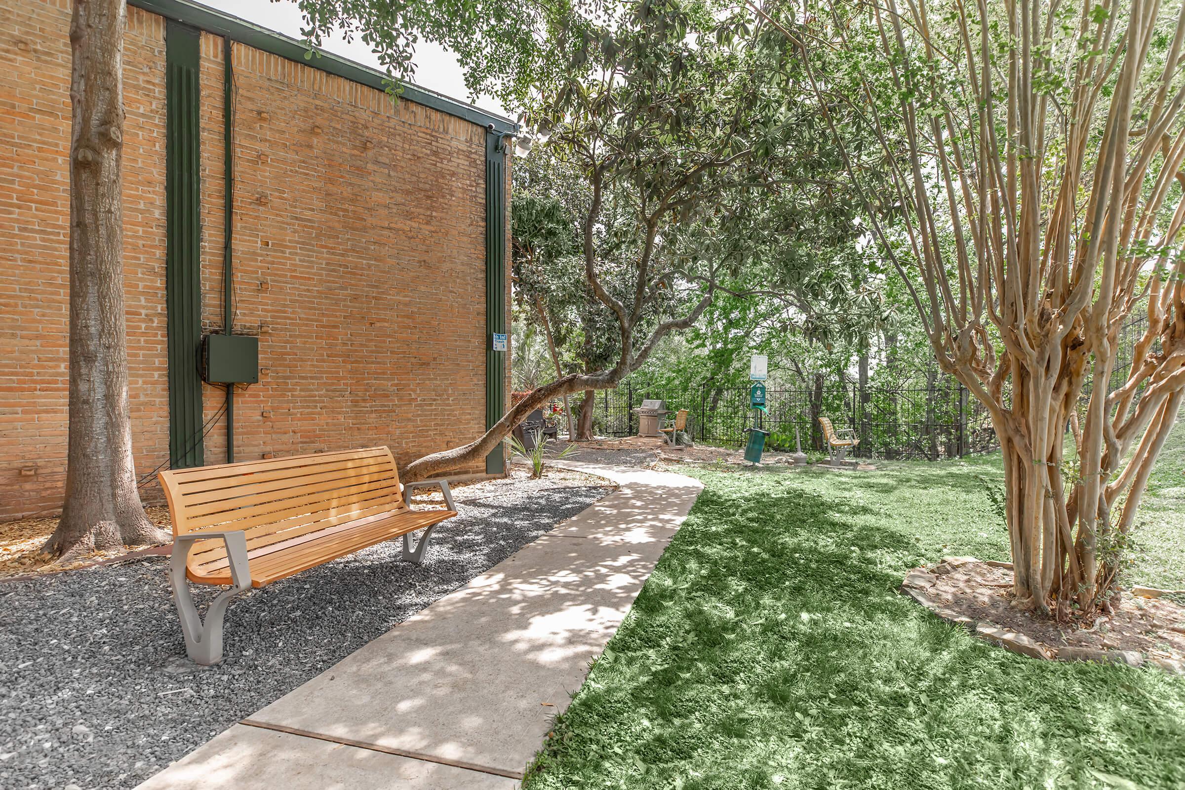 A wooden bench is positioned on a path surrounded by greenery and trees. A brick wall provides a backdrop, with a small utility box visible. The area features grass and a few objects in the background, creating a serene outdoor setting suitable for relaxation.