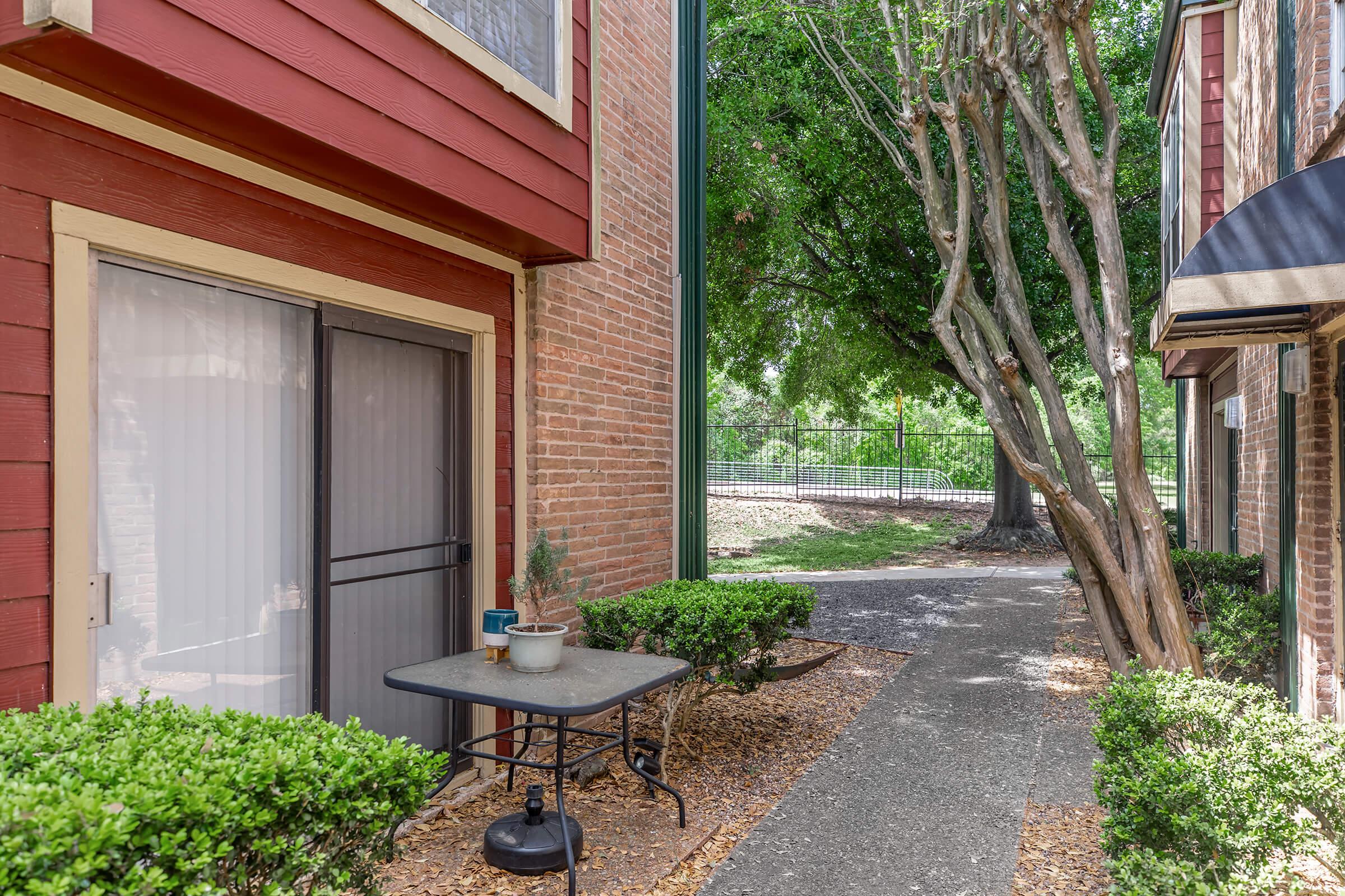 A small outdoor space featuring a table and potted plant beside a building. The area is bordered by neatly trimmed hedges and has a pathway leading to a gated area with trees in the background. The setting appears well-maintained and inviting.