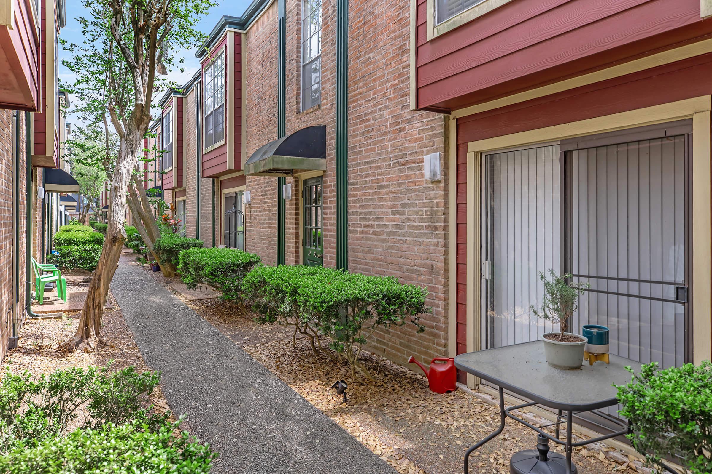 A narrow walkway flanked by brick apartment buildings, featuring manicured bushes and trees. Small tables with potted plants are visible, along with decorative awnings over the doors. Natural light illuminates the scene, creating a welcoming atmosphere.