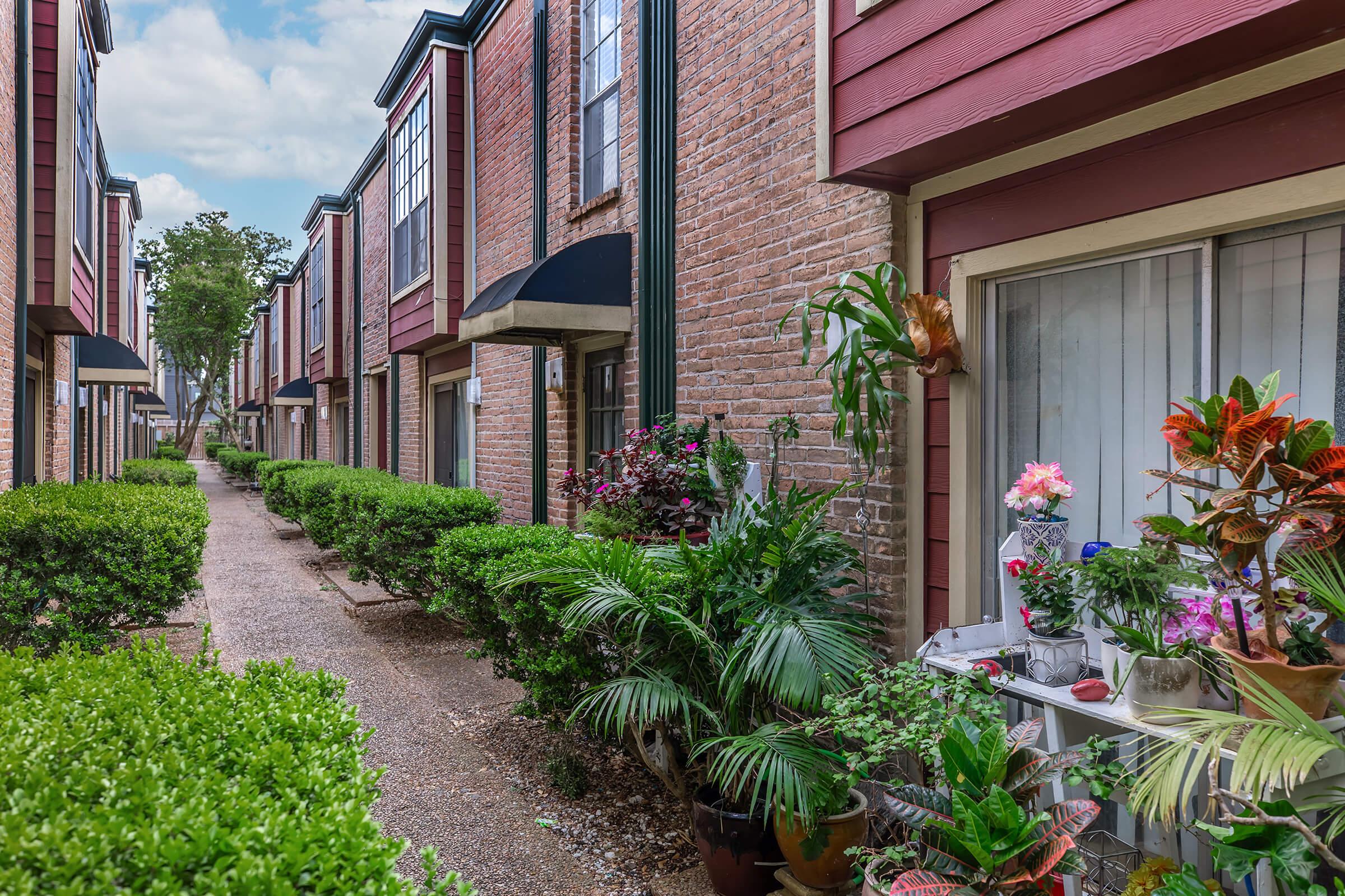 A peaceful walkway lined with manicured hedges and colorful plants, leading to brick buildings with red trim. Potted flowers and greenery adorn a small table on one side, while the path is illuminated by soft natural light, creating a serene outdoor atmosphere in a residential setting.