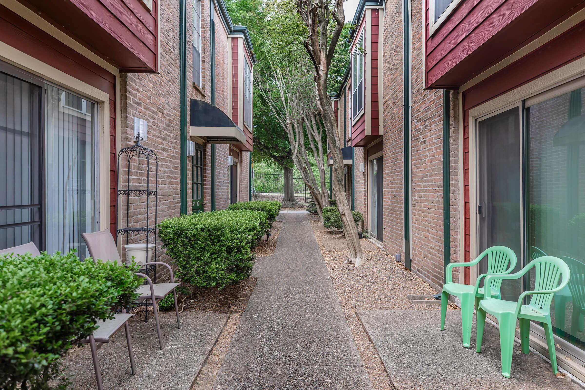 A narrow walkway between two brick buildings, lined with hedges. There are two green plastic chairs and a few metal chairs arranged on the sides. Trees and greenery can be seen in the background, creating a peaceful, shaded ambiance.