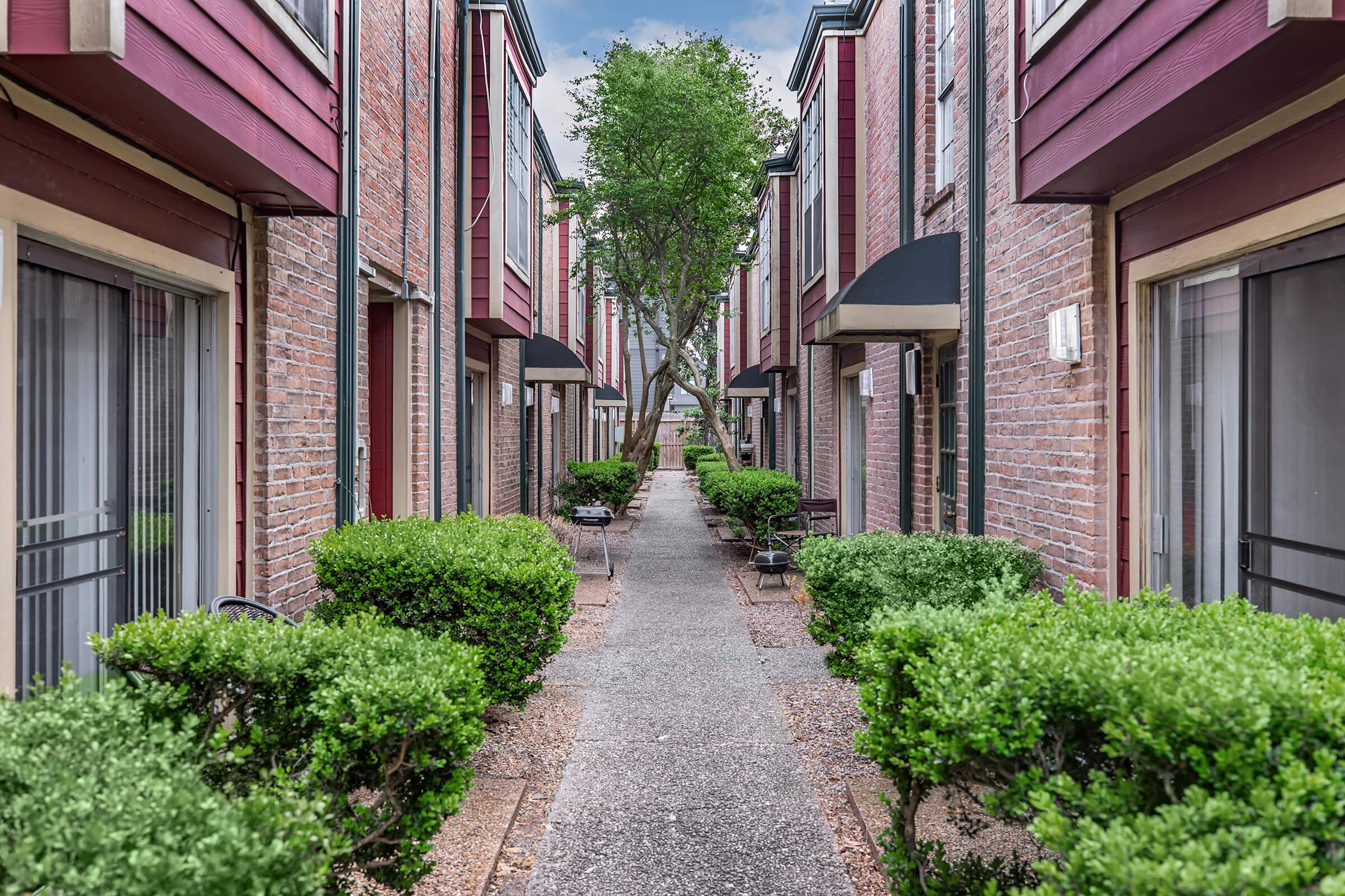 A narrow pathway lined with neatly trimmed hedges, leading between two rows of brick townhouses with red accents. Each unit features large windows and awnings, with a tree visible at the far end of the path, suggesting a well-maintained outdoor space.