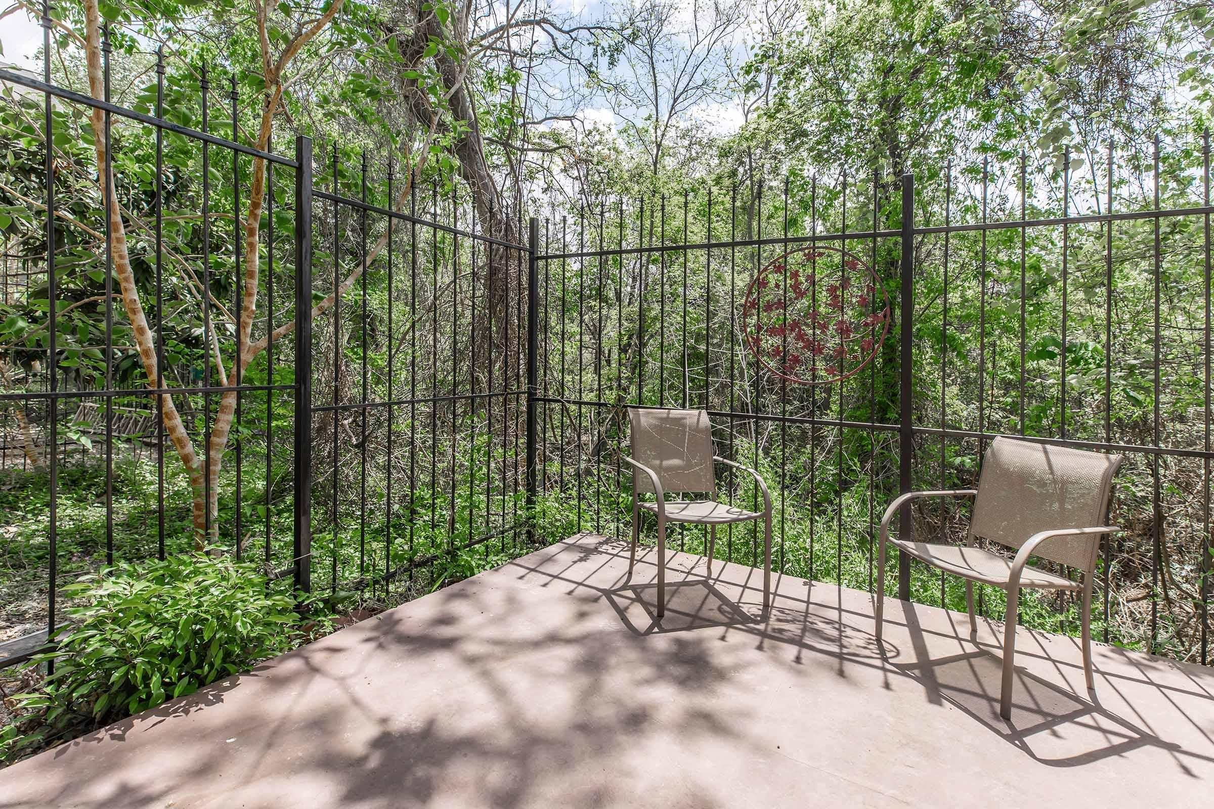 A small outdoor seating area featuring two metal chairs on a concrete patio, surrounded by a black metal fence. Lush greenery and trees are visible in the background, creating a serene and natural setting. Sunlight filters through the leaves, casting shadows on the ground.