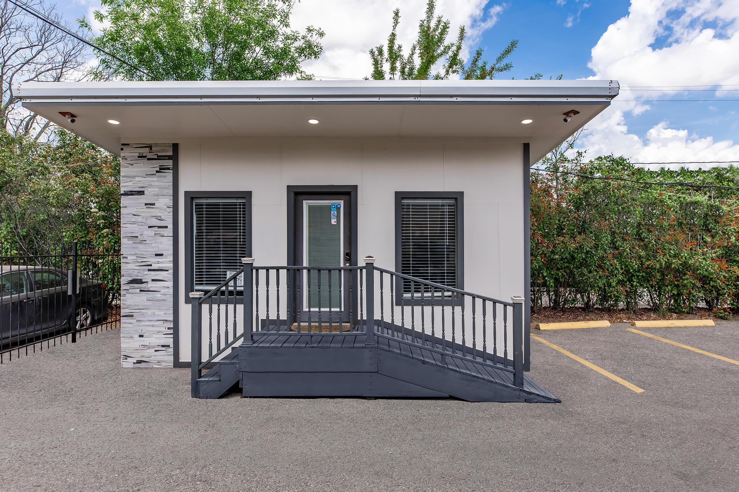 A modern single-story building with a stone accent wall and a wheelchair ramp at the entrance. It features large windows with dark frames and is surrounded by greenery. The sky is partly cloudy, and there are parked cars in the background. The overall setting appears inviting and accessible.