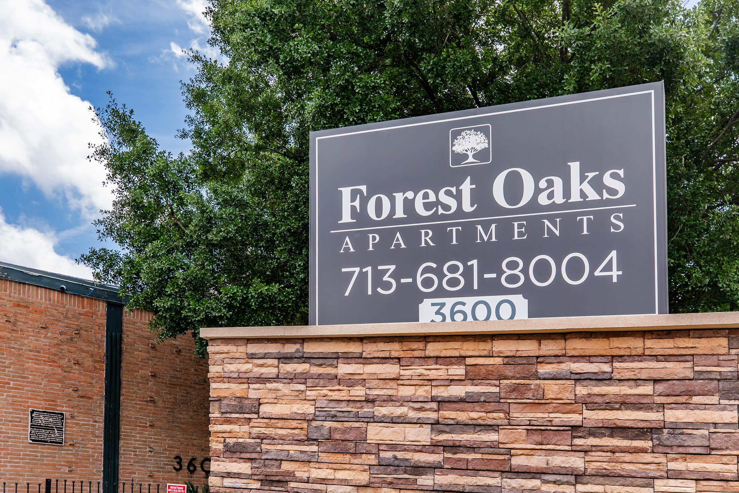 Sign for Forest Oaks Apartments featuring the name "Forest Oaks," a phone number (713-681-8004), and address (3600). The sign is set against a backdrop of greenery and a brick structure, with a clear blue sky and clouds overhead.