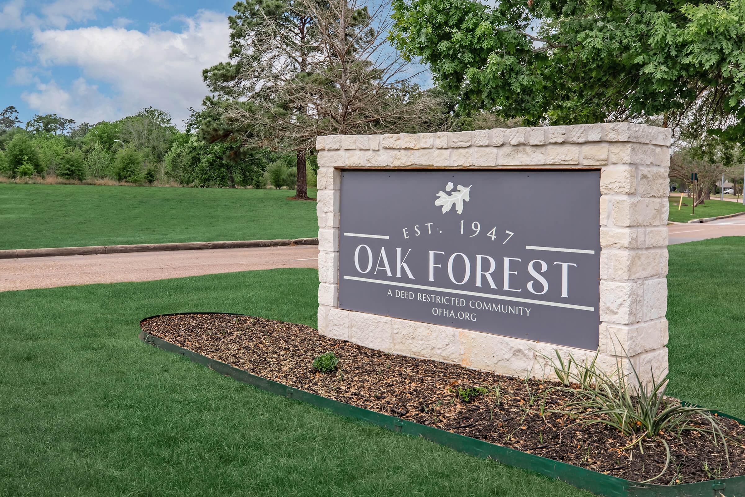 Sign for Oak Forest, established in 1947, indicating it is a deed-restricted community. The sign features a stone base and is positioned in a grassy area with trees in the background. The text is clearly visible, promoting the community's website, ofha.org.