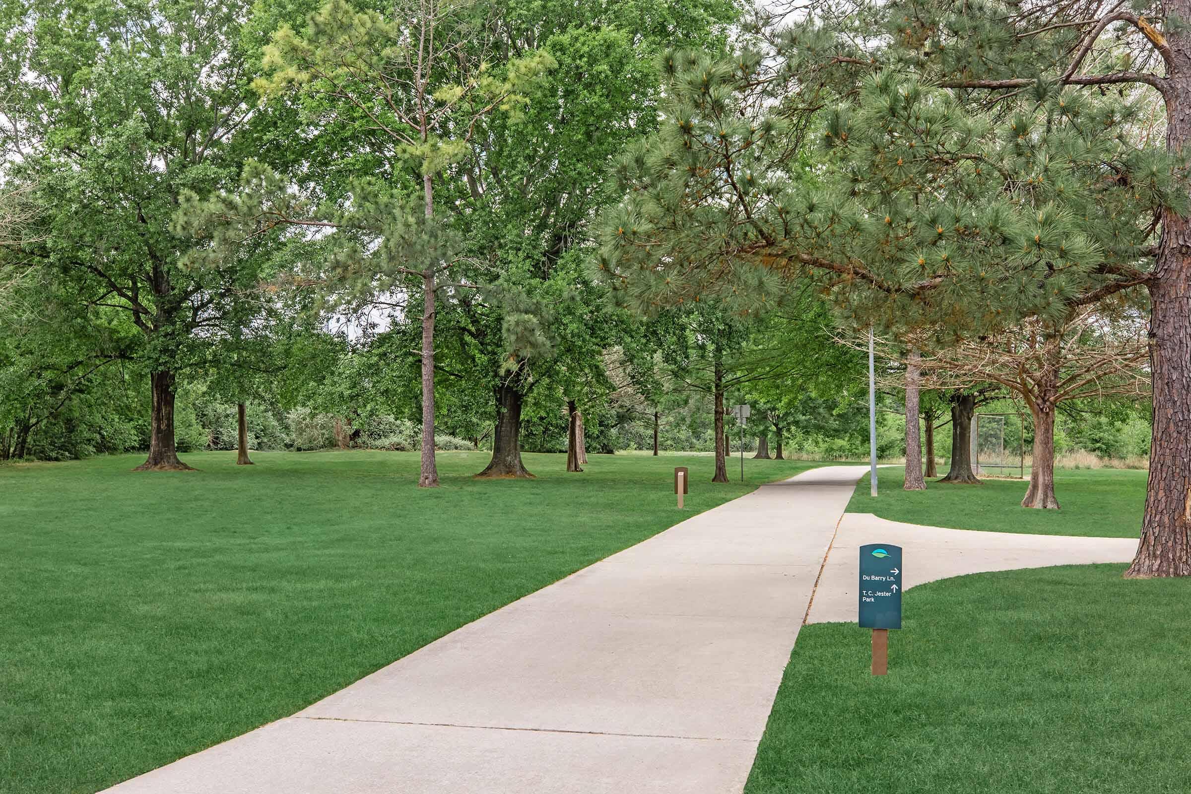 A scenic park pathway lined with tall trees and lush green grass. The paved path winds through the area, flanked by various trees for shade. A sign is partially visible on the right side of the path, indicating directions or information about the park. The setting appears peaceful and inviting for walking or relaxation.