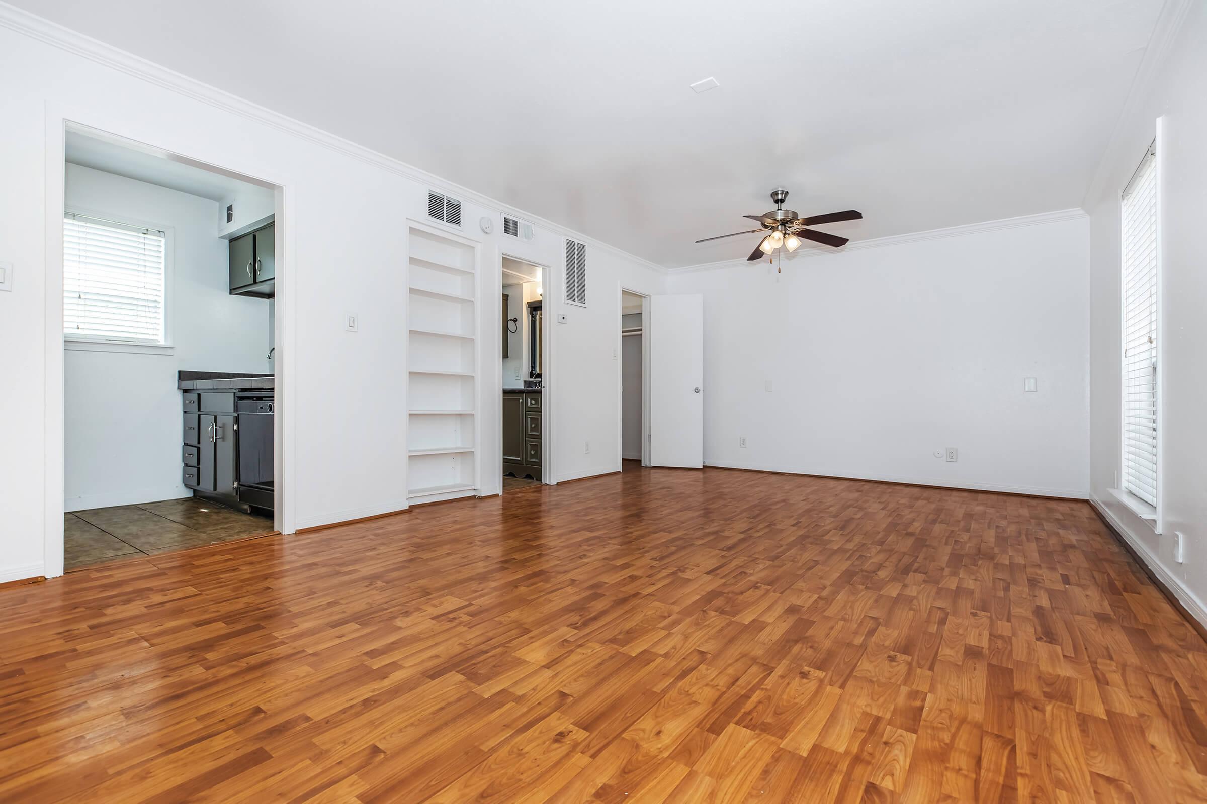 A spacious, well-lit living room featuring hardwood flooring, a ceiling fan, and white walls. An open concept connects to a kitchen area, visible through a doorway on the left. There are built-in shelves along one wall and a door leading to another room on the right. Natural light comes from a window, enhancing the airy feel.