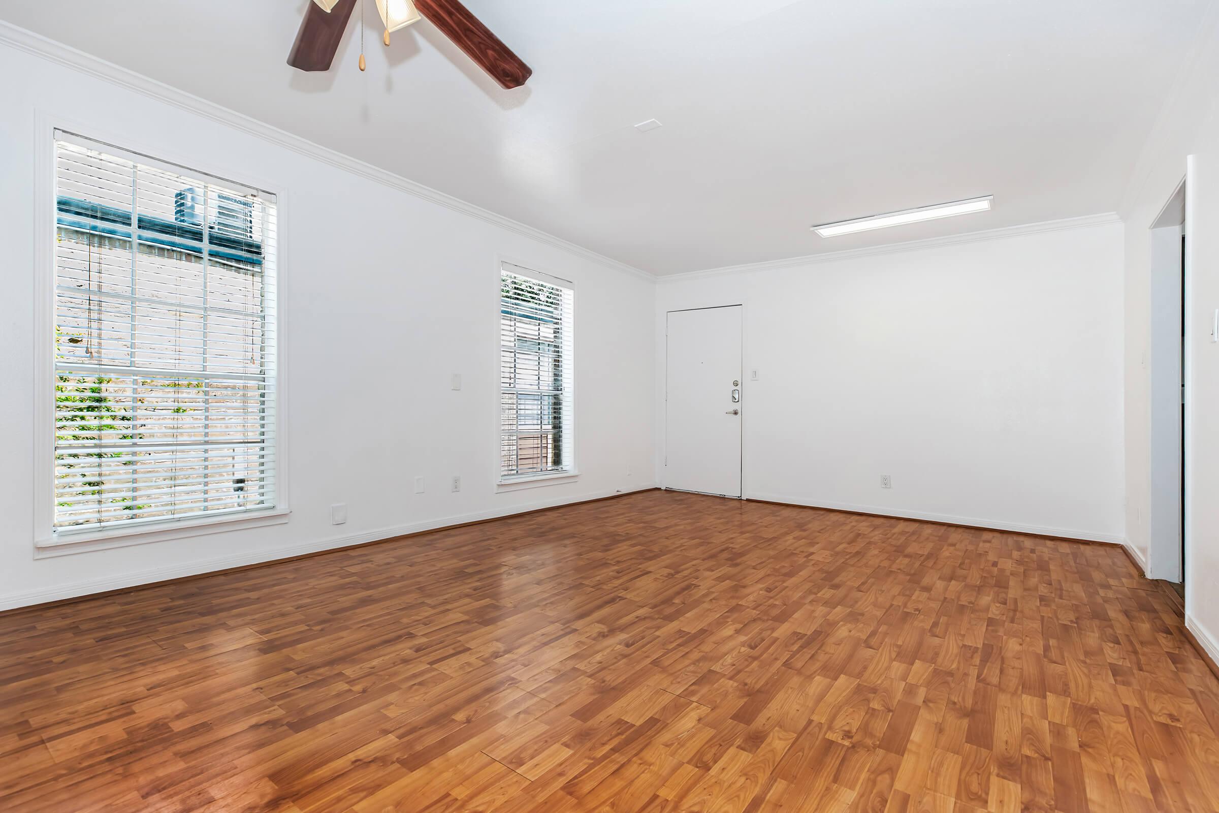 A bright, empty room with hardwood flooring, featuring a ceiling fan and large windows with blinds. The walls are painted white, and there is a door on one side. Natural light fills the space, creating an airy atmosphere.