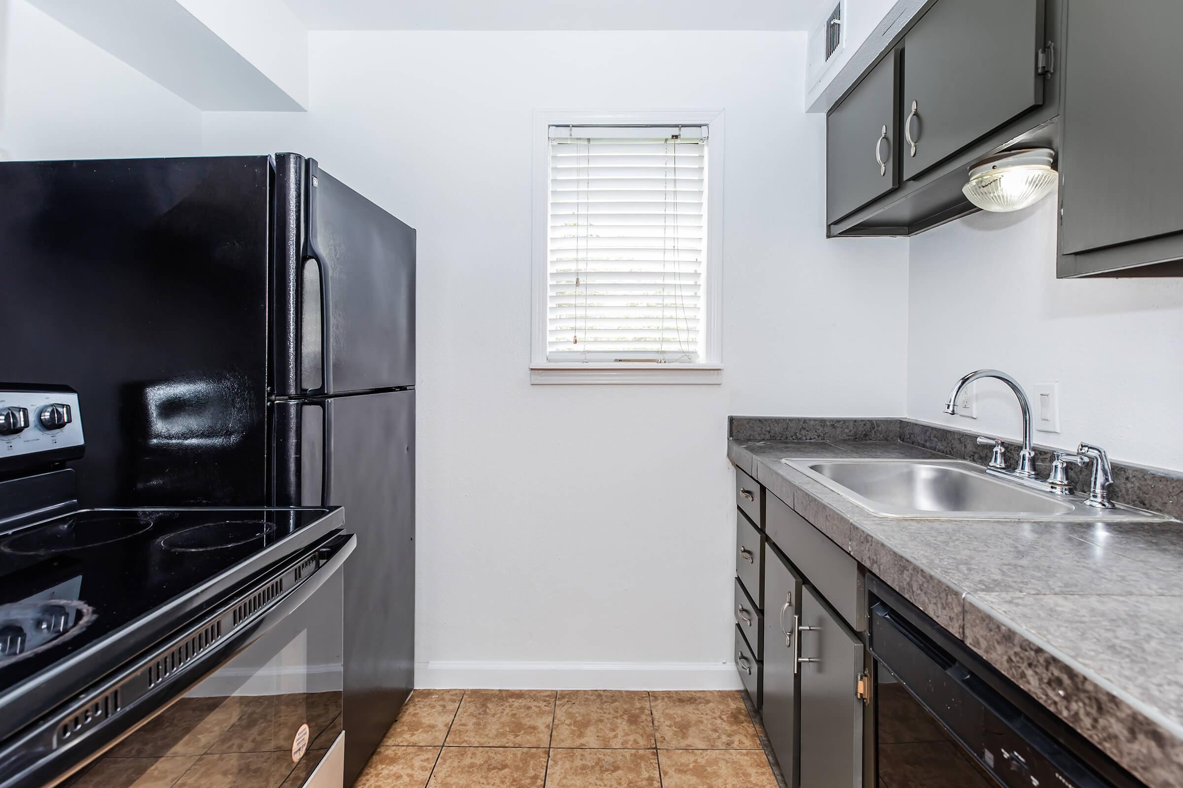 Modern kitchen featuring black appliances, including a refrigerator and stove, with gray cabinets and countertops. A window with blinds allows natural light, and the floor is tiled in a light brown pattern. Overall, the space appears clean and functional.