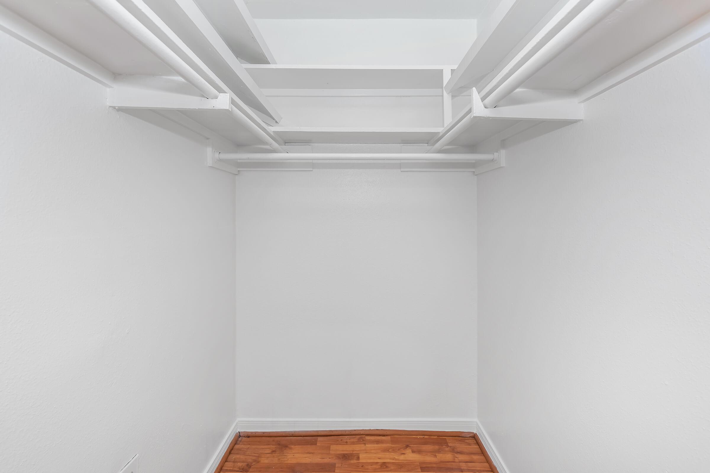 A spacious, empty closet with white walls and a wooden floor. The closet features white shelving and hanging rods along the top, providing ample storage space. The overall look is clean and minimalist, suitable for organizing clothes or other items.