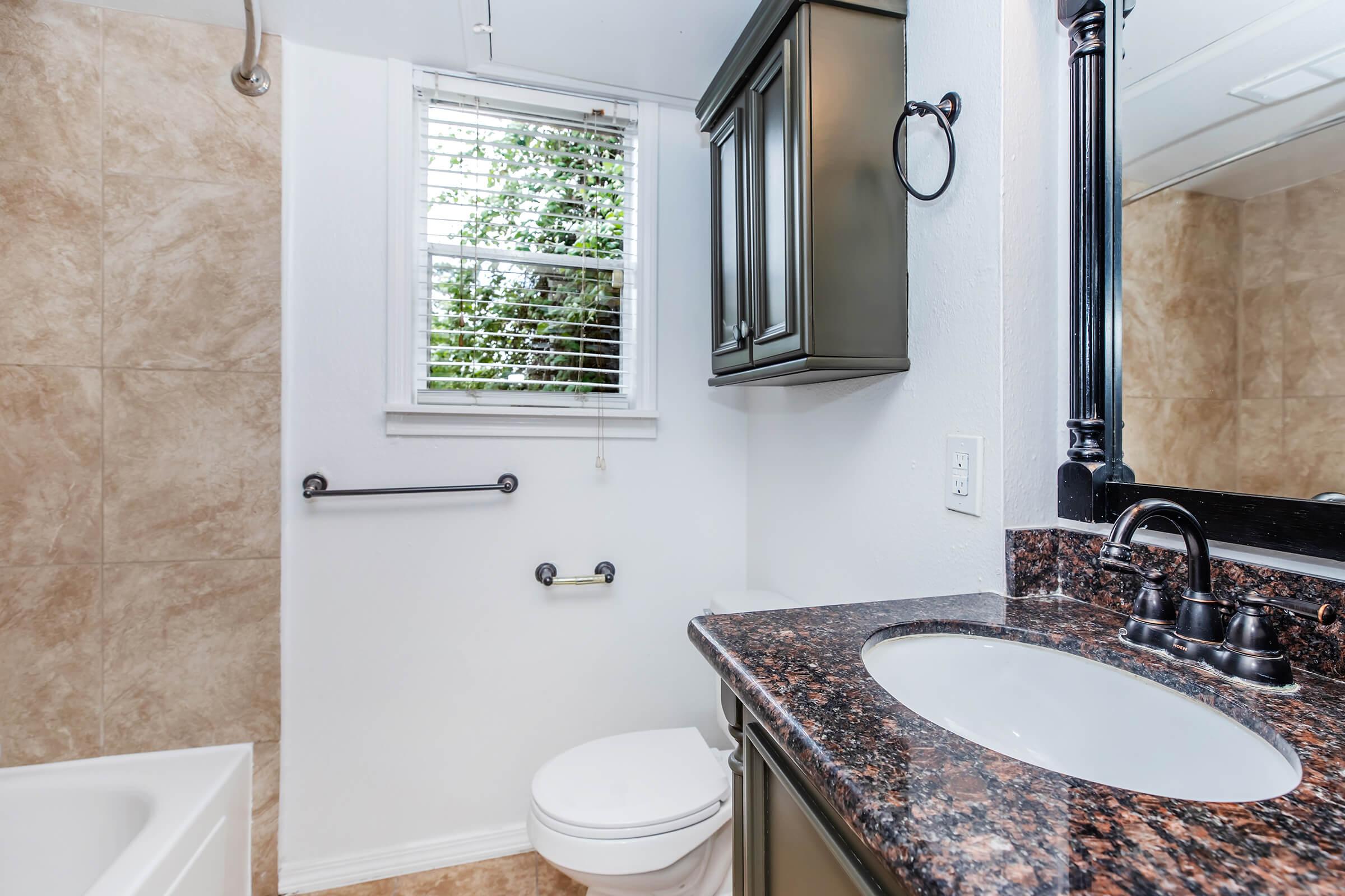 A modern bathroom featuring a white toilet, a granite countertop sink, and a large mirror. There's a window with blinds allowing natural light, and a bathtub is visible in the background. The walls are tiled with a neutral tone, and there are chrome fixtures.