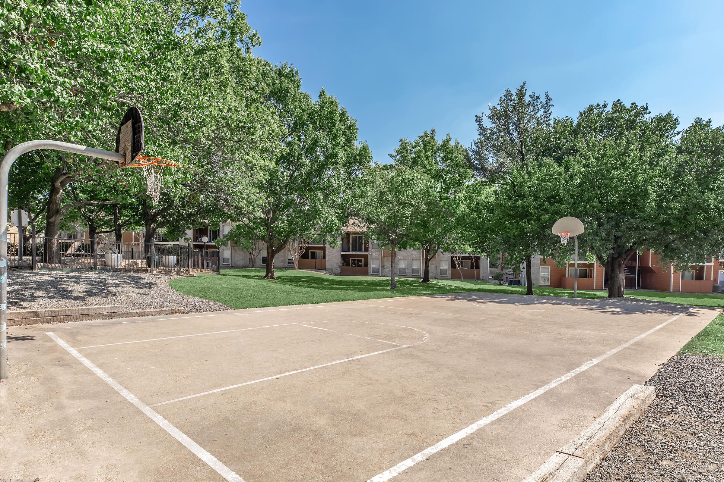 Outdoor basketball court with a hoop and backboard, surrounded by lush green trees and grass. The court features a smooth, concrete surface with a painted boundary. In the background, there are residential buildings with large windows, creating a serene, recreational environment.