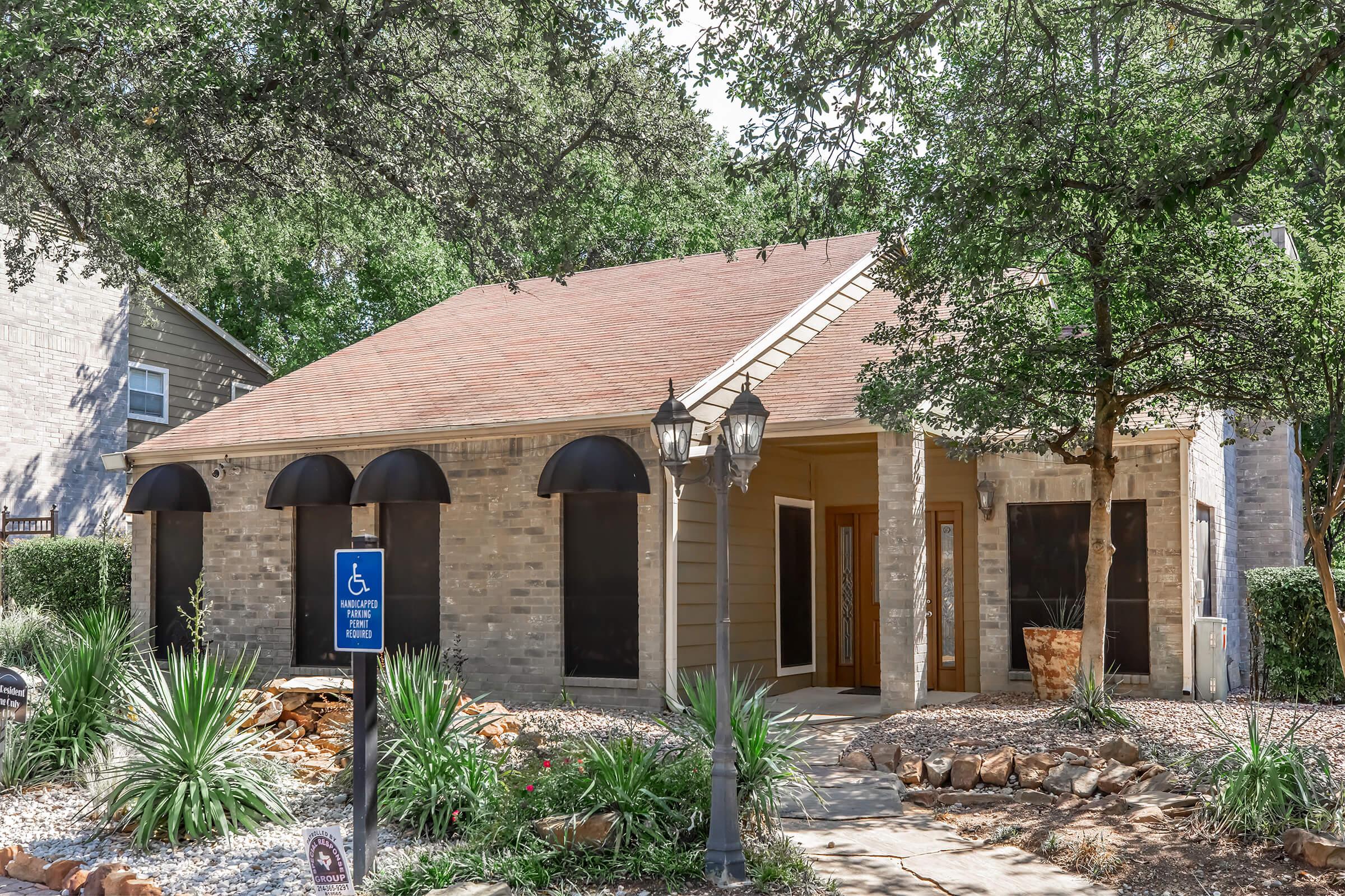 A single-story house with a tan exterior, featuring black awnings above the windows. The front yard includes rocks, shrubs, and a wheelchair accessible sign. Decorative lanterns flank the walkway leading to the entrance, surrounded by trees and other greenery.