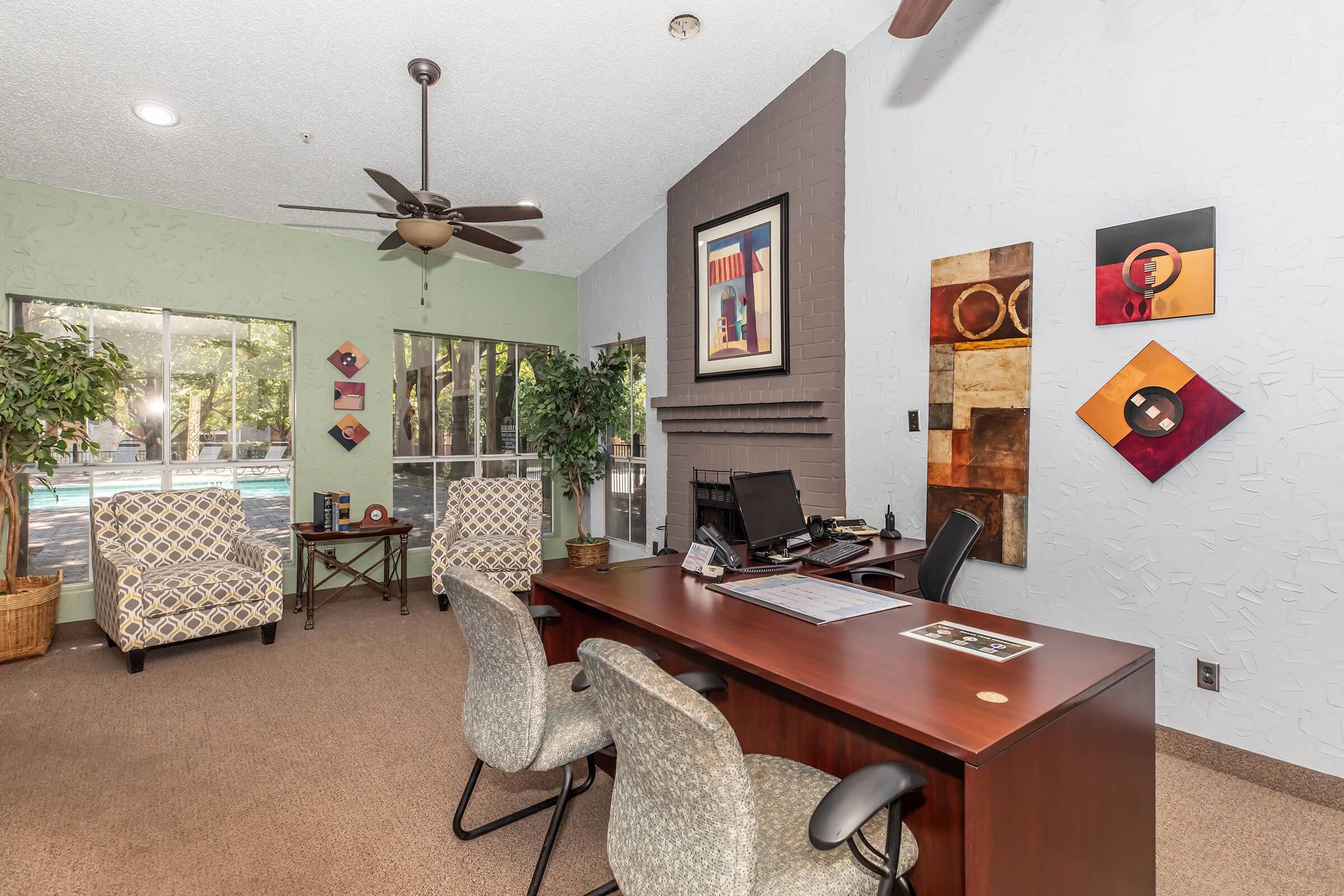 A well-lit office space featuring a wooden desk with a chair, surrounded by two patterned armchairs and indoor plants. The walls are adorned with framed artwork, and large windows provide a view of an outdoor pool area. A ceiling fan is visible, adding to the inviting atmosphere.