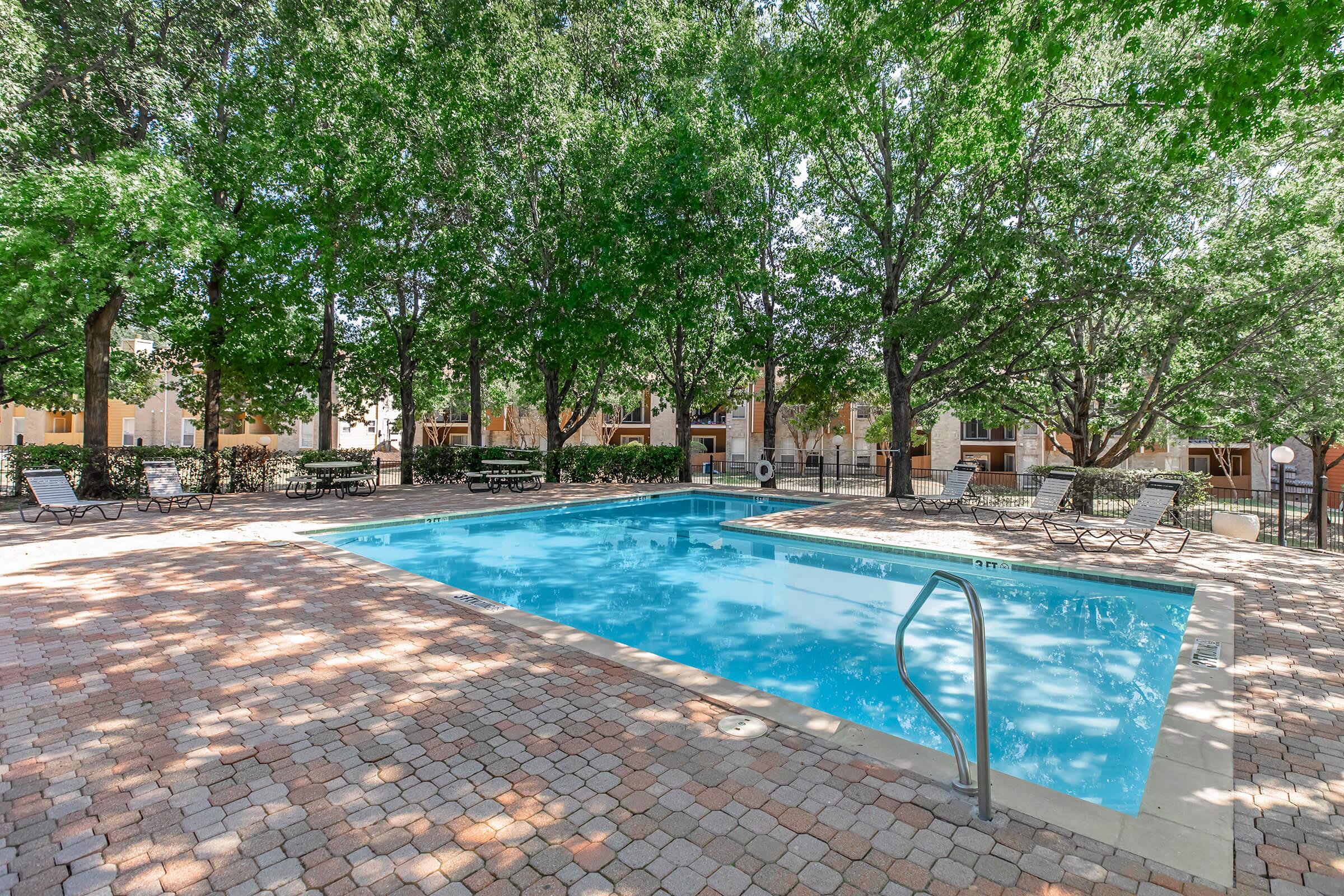 A clear swimming pool surrounded by lush greenery and trees. Lounge chairs are set around the pool area, with a brick-paved deck. The scene is bright and inviting, suggesting a relaxing outdoor space for leisure and recreation.