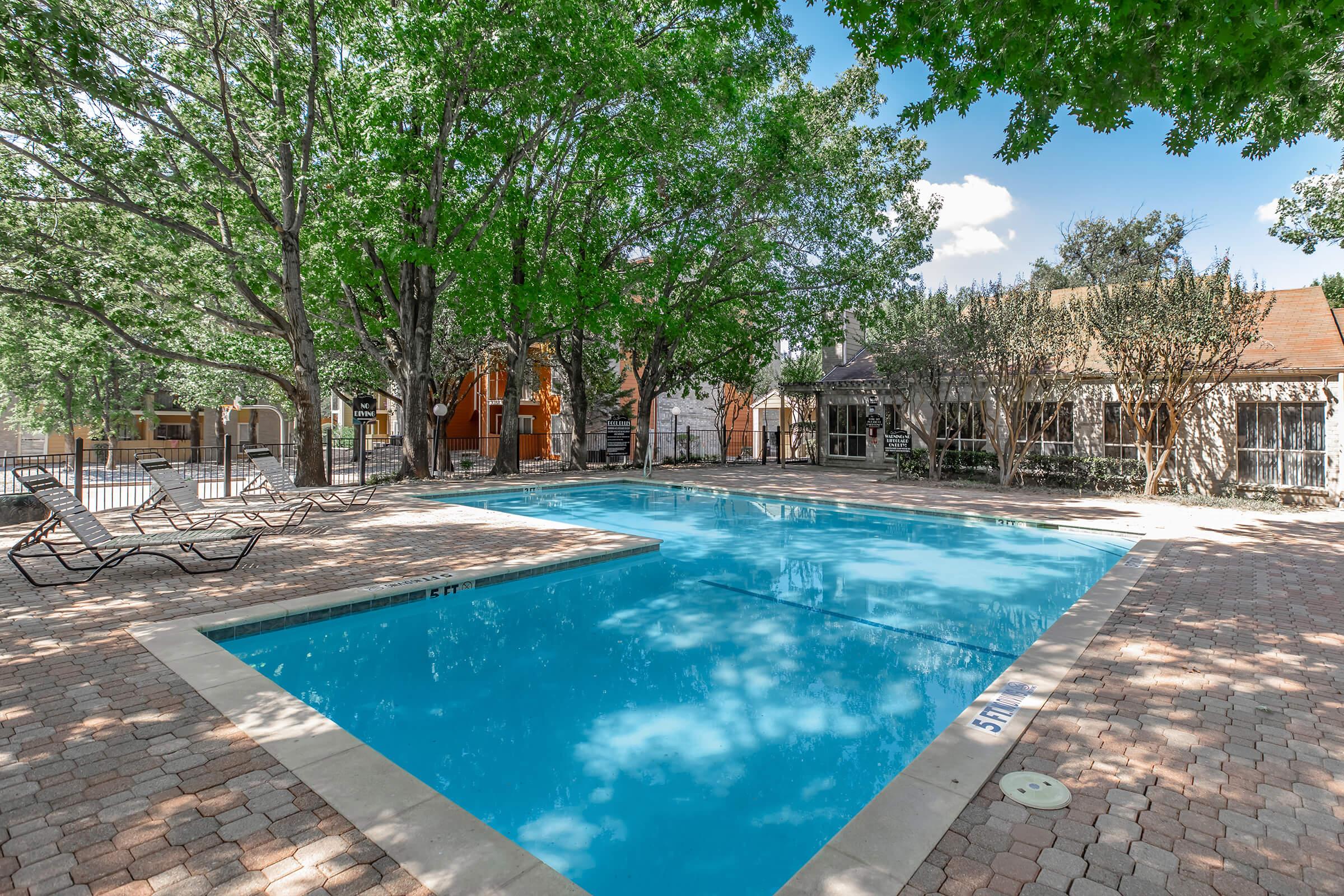 A bright, inviting swimming pool surrounded by lush green trees and neatly arranged lounge chairs. The pool is set within a landscaped area, featuring brick paving, with sunlit reflections on the water's surface and a few buildings in the background. Ideal for relaxation and recreation.