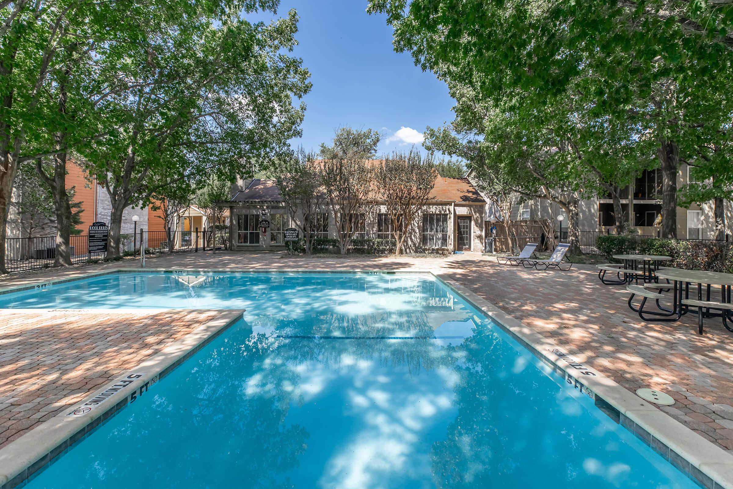 A serene outdoor swimming pool surrounded by greenery and trees, with lounge chairs and picnic tables nearby. In the background, there is a building with large windows, and the sky is partly cloudy. The pool area is well-maintained, inviting relaxation and leisure.