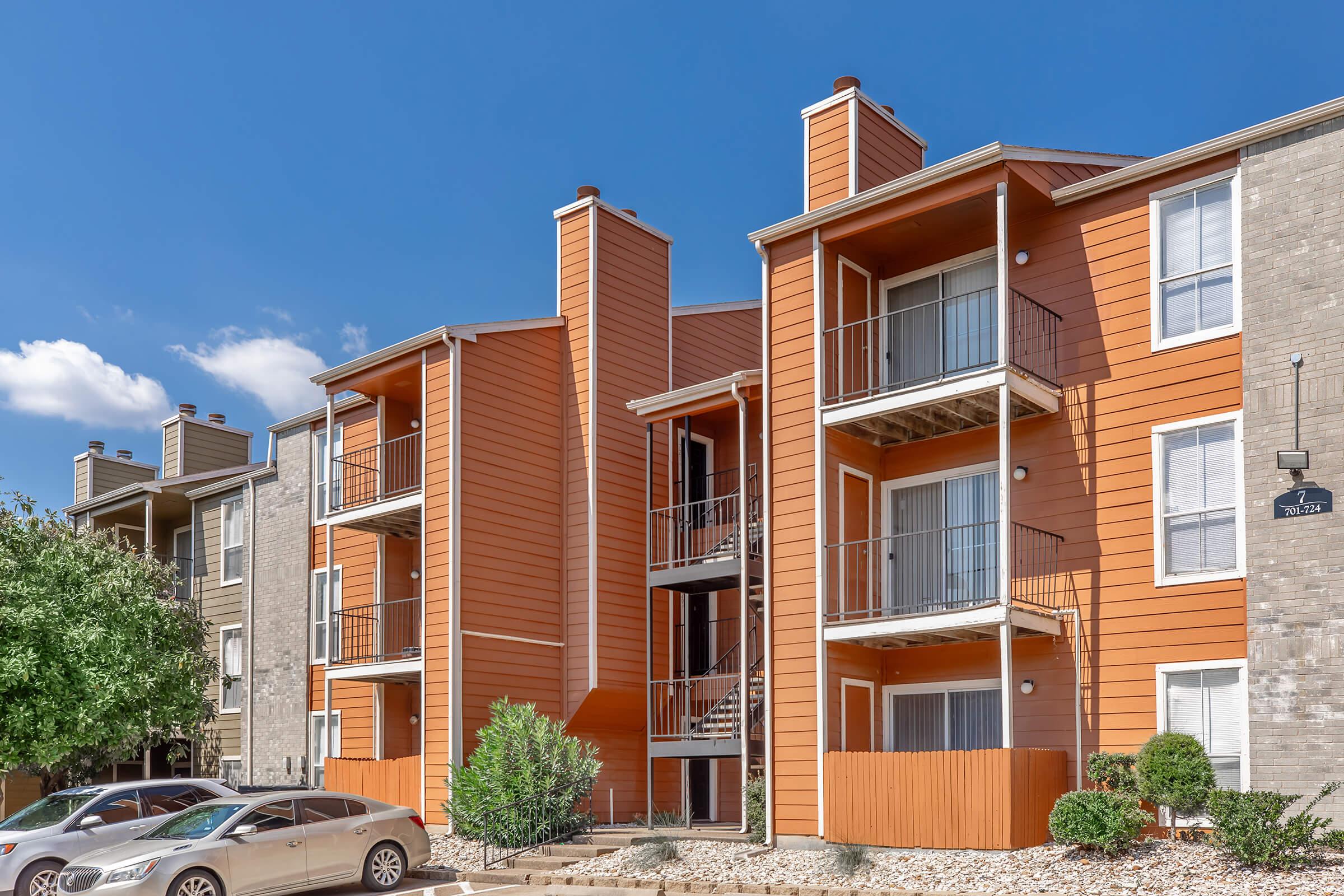 Apartment building exterior featuring multiple stories with orange and tan siding. Balconies and staircases are visible, along with a landscaped area and parked cars in the foreground. The sky is clear with a few clouds, creating a bright atmosphere.