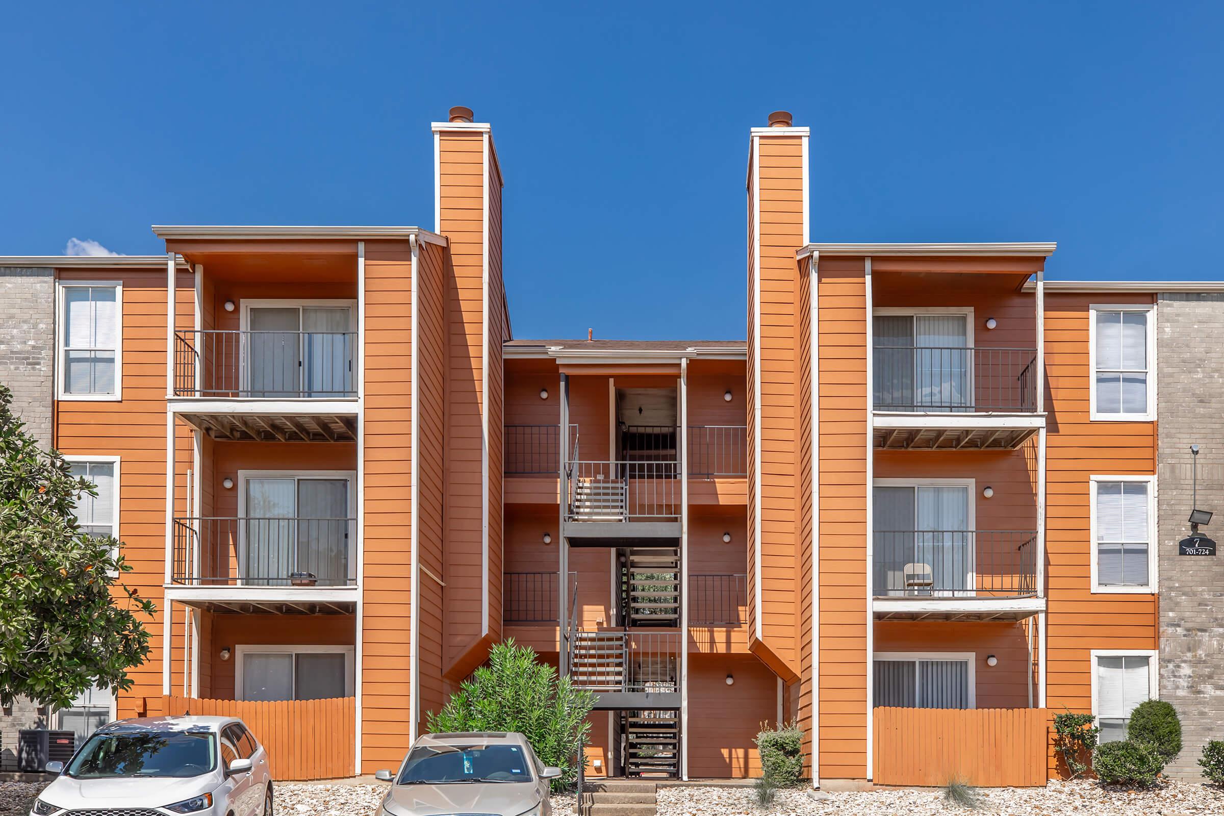 A multi-story apartment building featuring orange and beige exteriors. The building has balconies with metal railings, a central stairway, and surrounded by small trees and shrubs. Two cars are parked in front, and the sky is clear and blue.