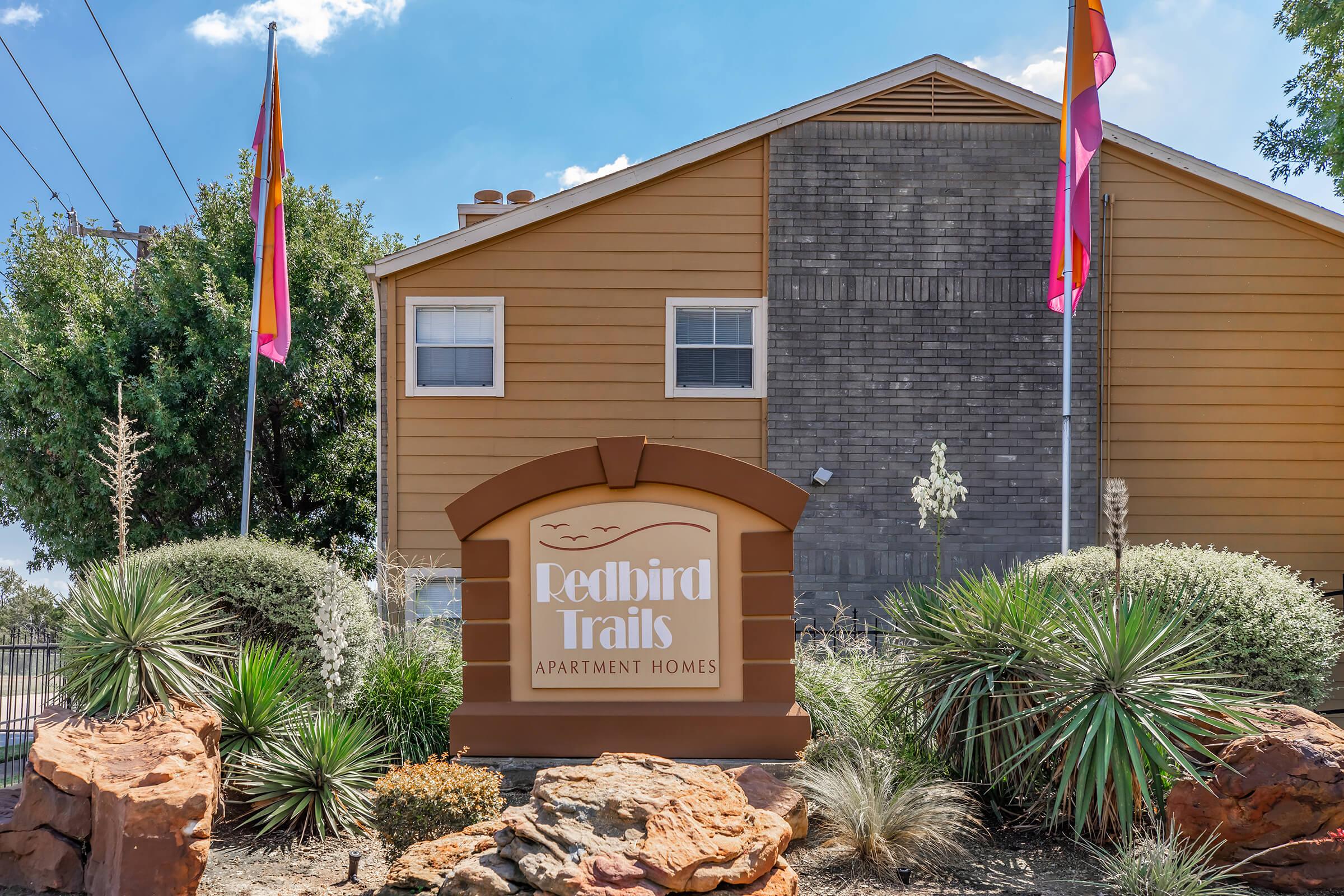 Sign for Redbird Trails Apartment Homes, featuring a decorative archway and landscaping with desert plants. The building behind is two stories with a light brown exterior, and flags are displayed on either side of the sign. The sky is clear with a few clouds.