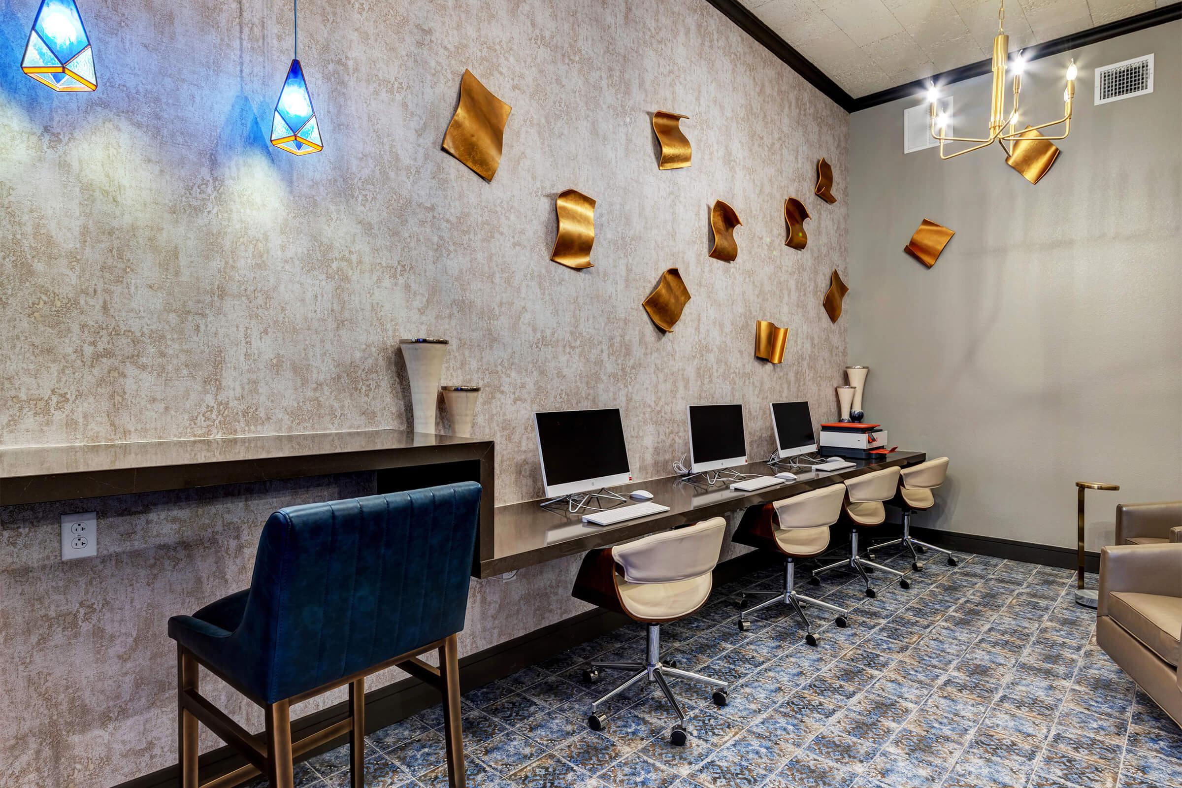 A modern workspace featuring several computer desks with sleek monitors, a light-colored wall adorned with decorative gold accents, and a stylish blue bar chair. The floor has a patterned tile design and there are pendant lights hanging from the ceiling, creating a contemporary and inviting atmosphere.
