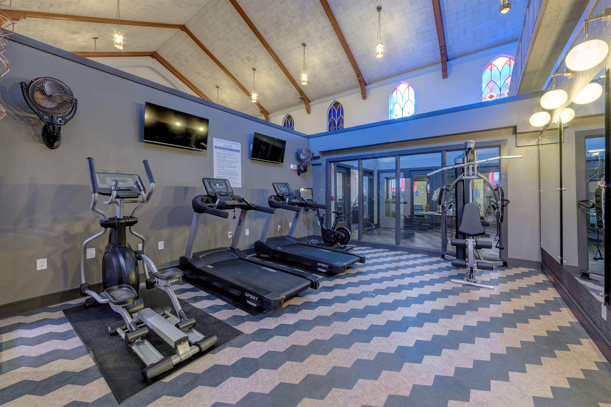 A modern gym interior with fitness equipment including treadmills, an elliptical machine, and a multi-station weight machine. The space features a unique design with high ceilings, decorative windows, and a stylish patterned floor, creating a bright and inviting workout environment.