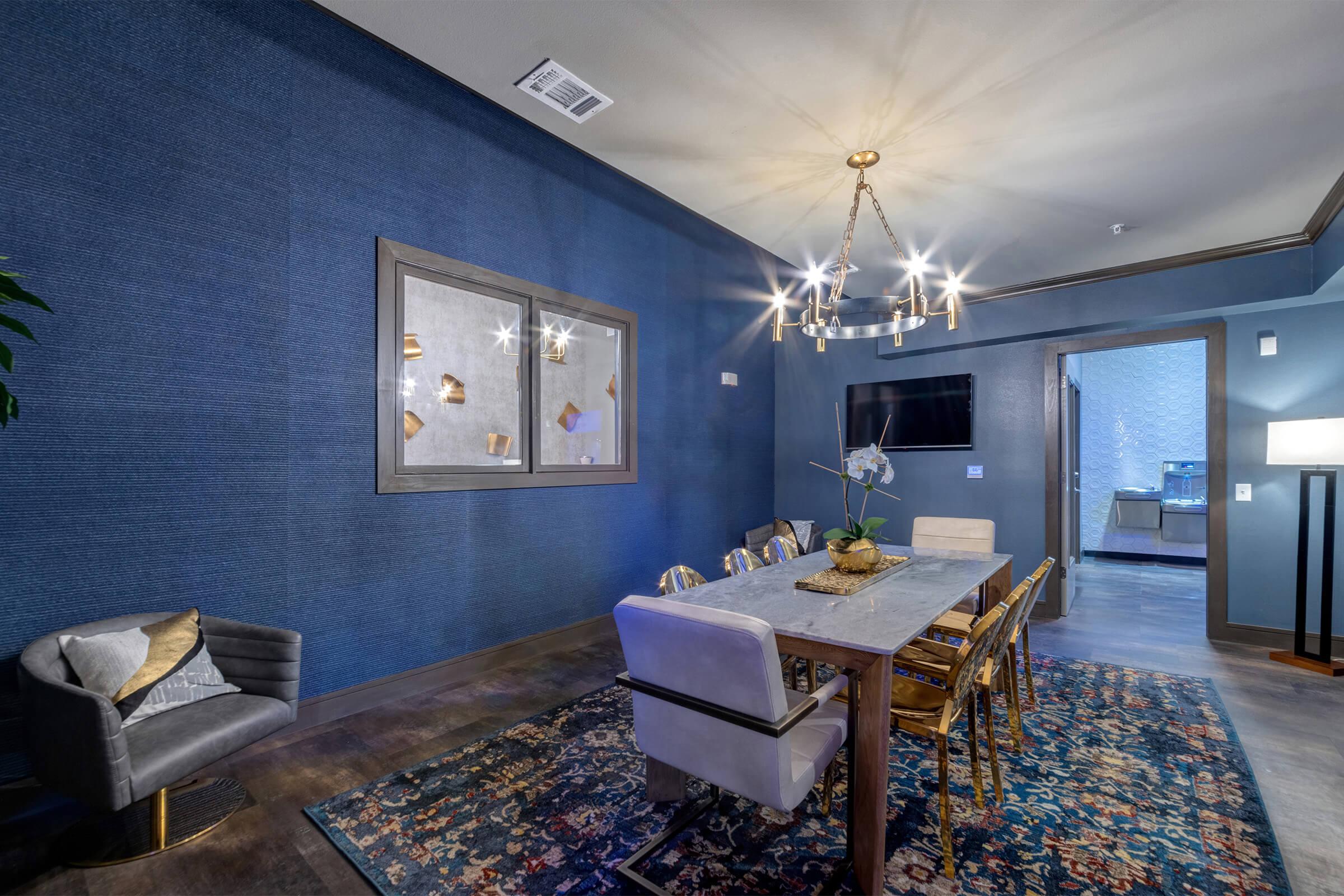 A stylish dining area featuring a long table with white chairs, a decorative rug, and a modern chandelier. The walls are deep blue, with a large framed mirror and a flat-screen TV. A potted plant adds a touch of greenery, and a doorway in the background leads to another room.