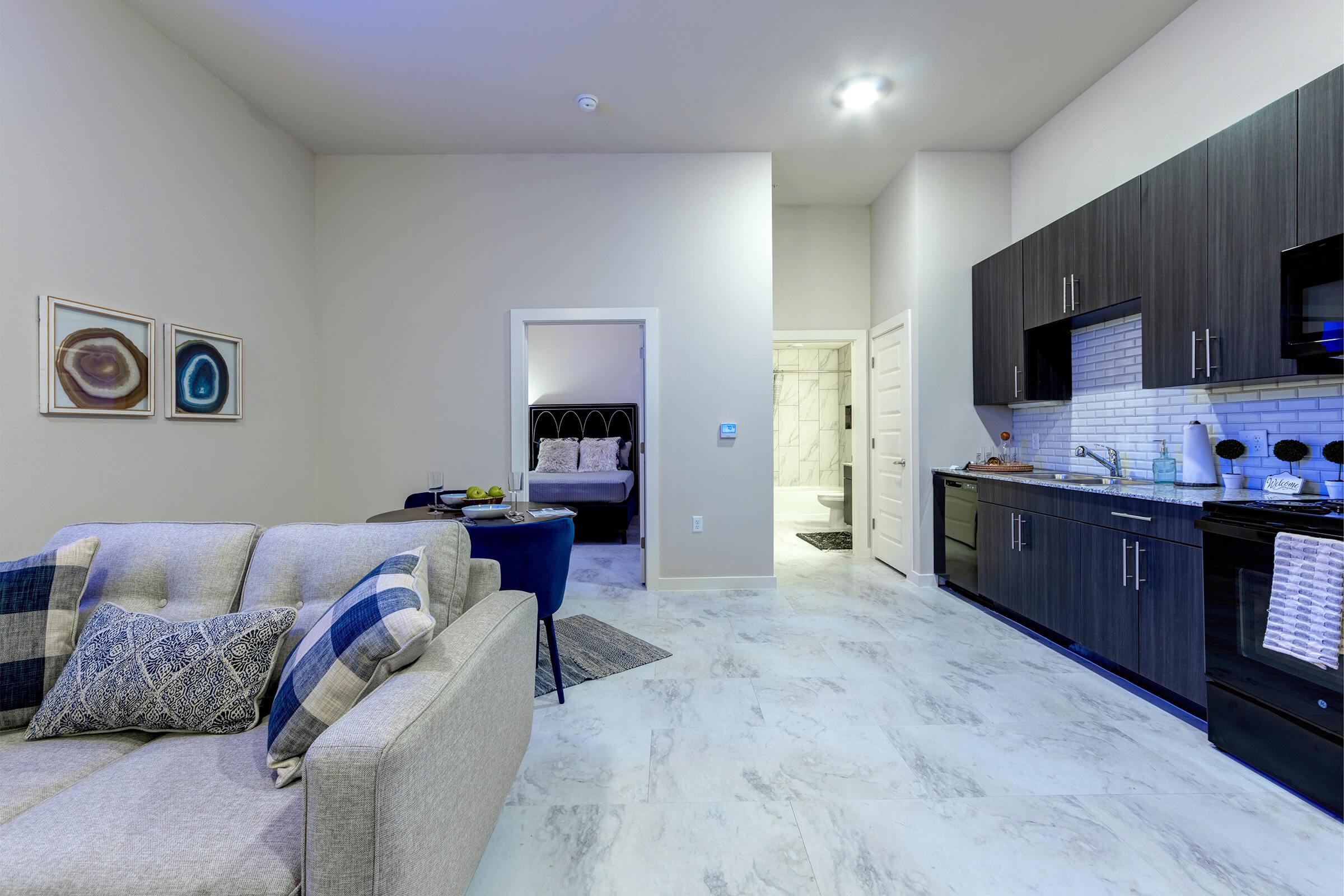 Interior of a modern apartment featuring a light-colored living area with a plush sofa, stylish wall art, and an open kitchen with dark cabinets. In the background, a bedroom is visible, along with a spacious bathroom entrance. The space is well-lit with warm lighting and has a clean, contemporary design.