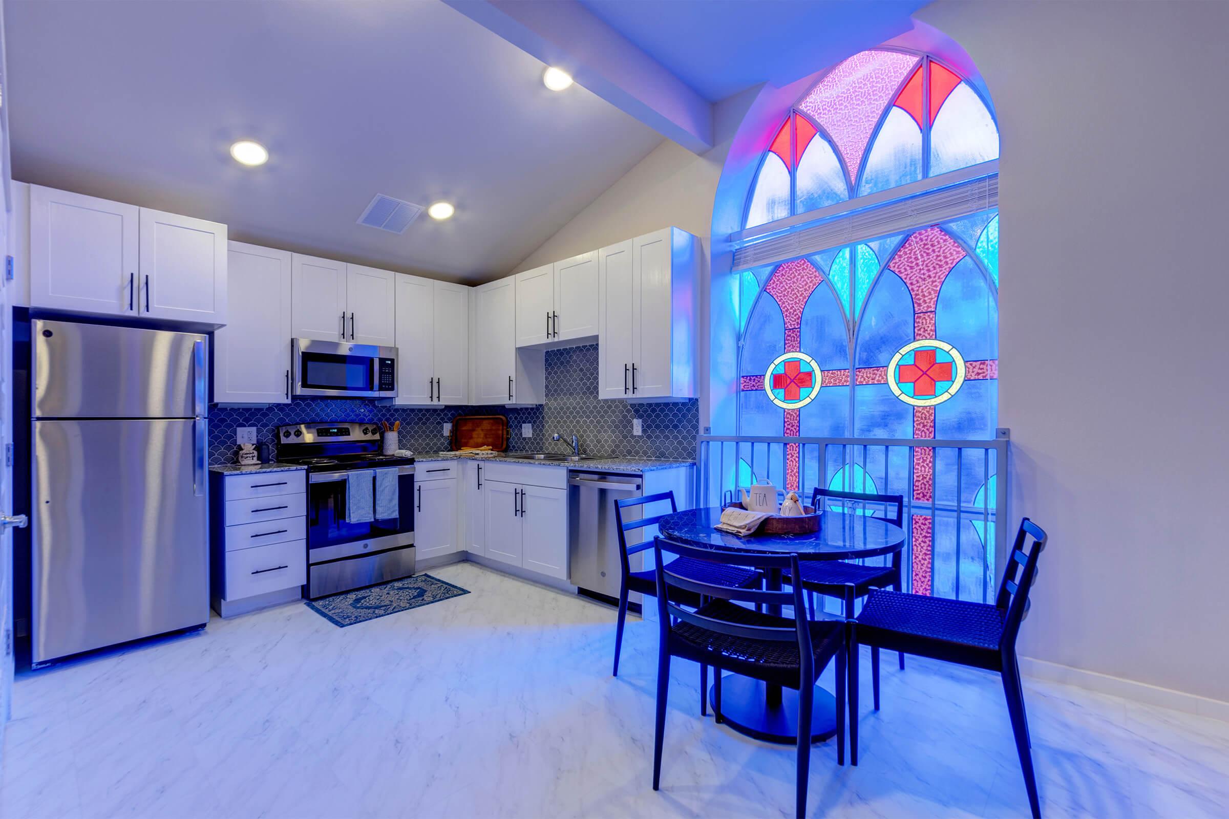 A modern kitchen featuring stainless steel appliances, white cabinetry, and a patterned backsplash. A round dining table with black chairs is positioned near a large, colorful stained glass window that adds a vibrant touch to the space. The floor is light-colored, enhancing the airy feel.