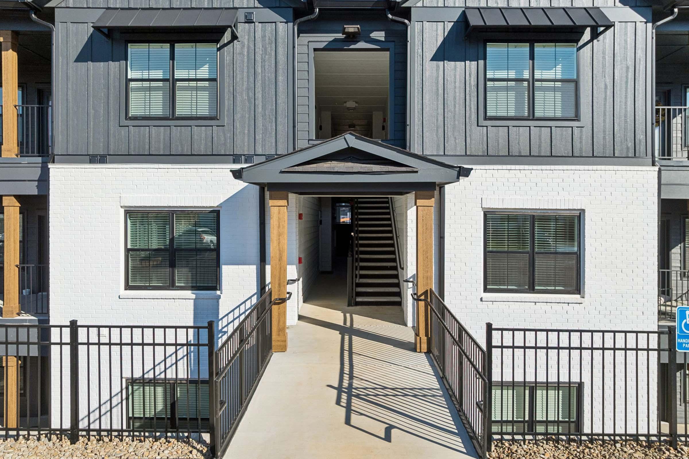 Entrance of a modern apartment building featuring a symmetrical façade with dark gray and white walls. A covered entryway leads to a set of stairs, flanked by large windows on either side. A black metal fence borders the walkway, which is accessible for individuals with disabilities.