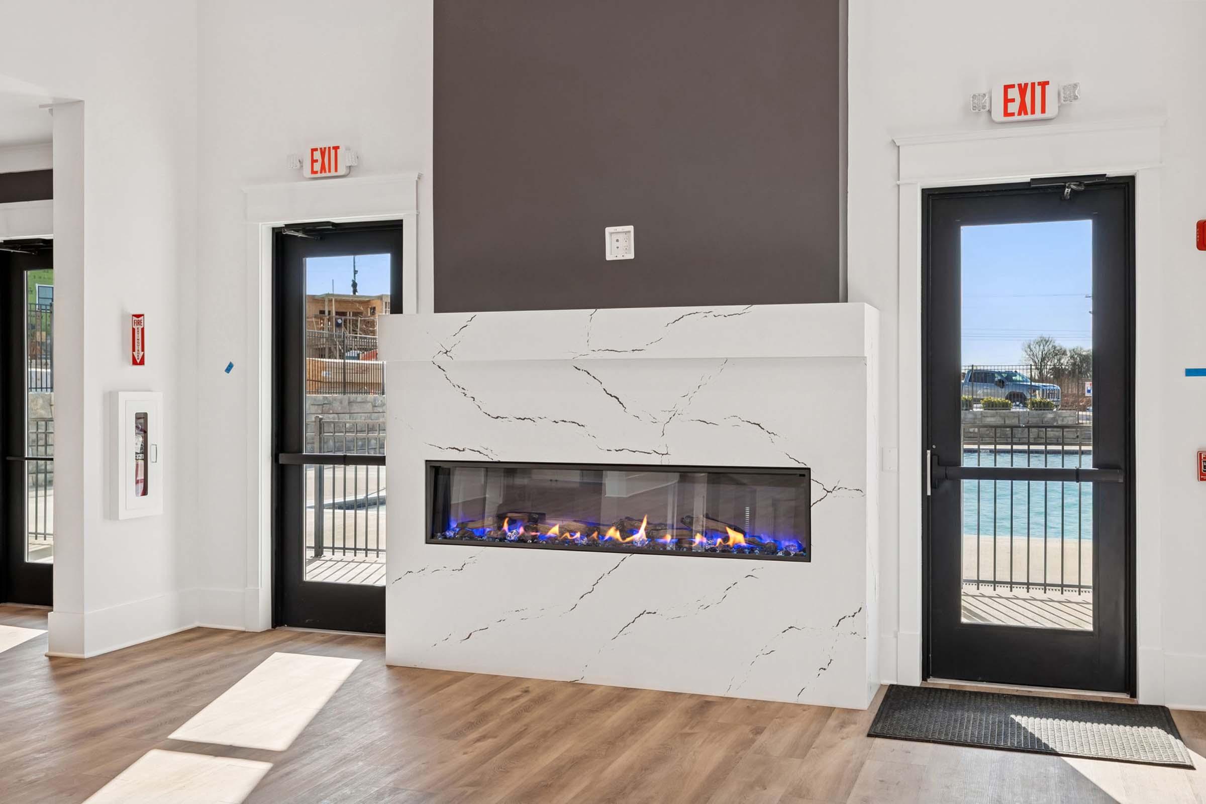 Modern interior featuring a sleek fireplace with blue flames, set against a white and gray wall. Two exit doors flank the fireplace, adding to the contemporary design. Natural light streams in from the windows, highlighting the flooring and creating a warm ambiance in the space.