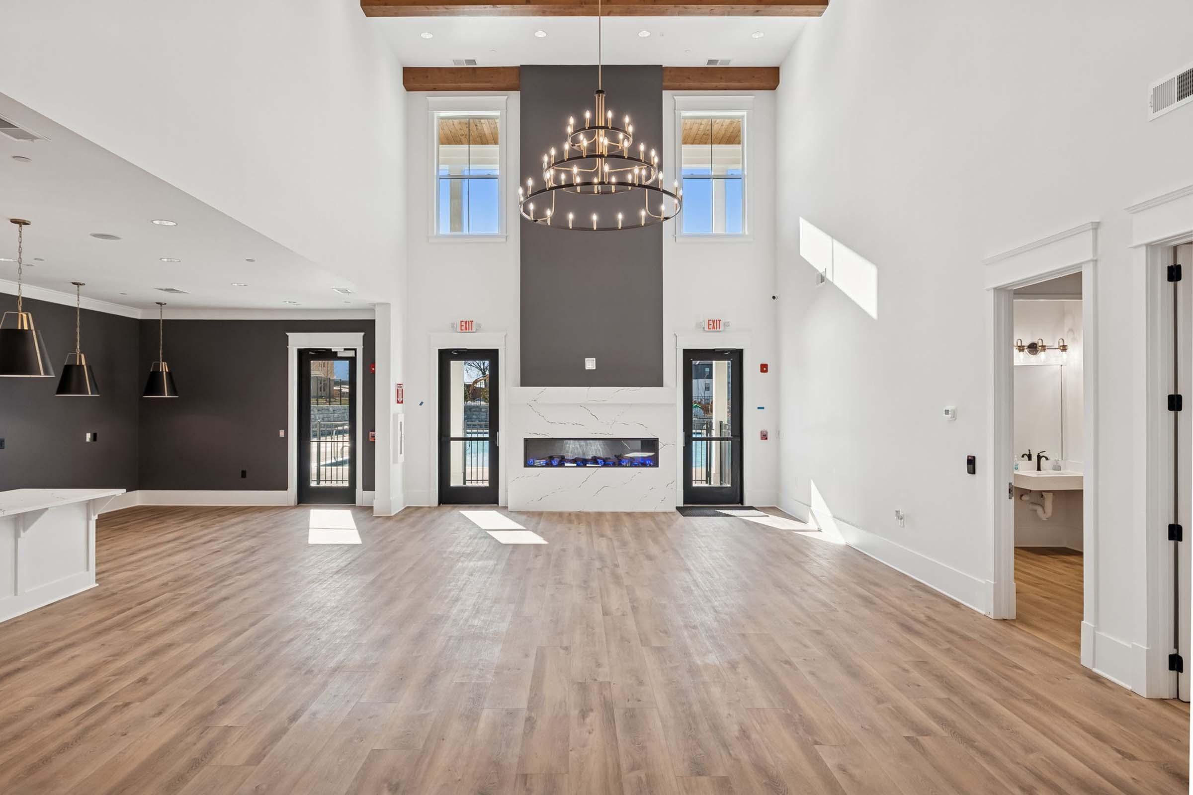 A spacious, modern interior featuring wooden beams, large windows providing natural light, and a stylish chandelier. The room has a light wooden floor and is decorated in neutral tones with a sleek fireplace. The layout suggests a welcoming atmosphere, suitable for gatherings or events.