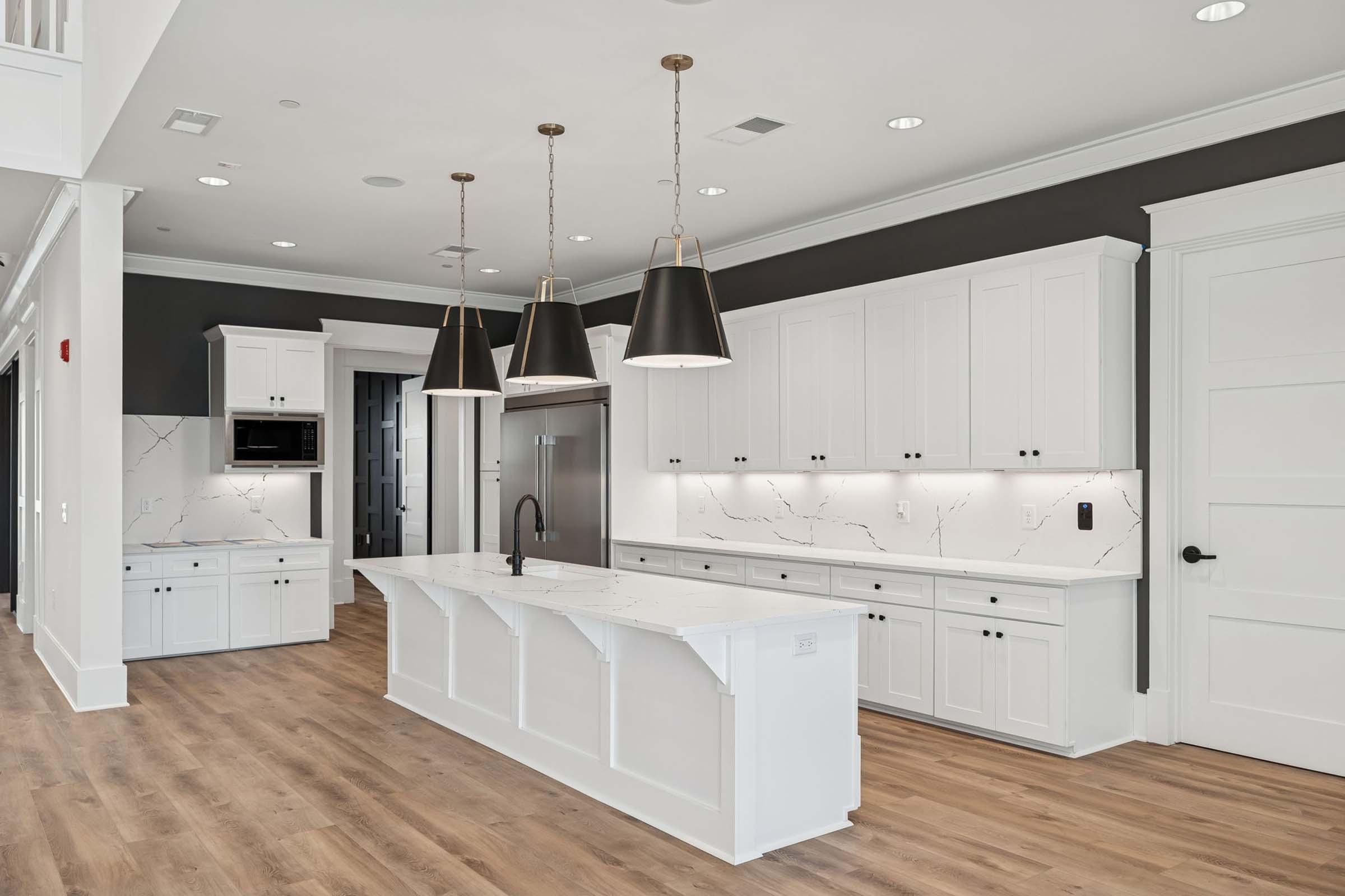 A modern kitchen featuring white cabinetry, a large central island with a marble countertop, and three pendant lights hanging above. The walls are a dark shade, contrasting with the light cabinetry. There is stainless steel appliances, including a refrigerator, and wooden flooring throughout.
